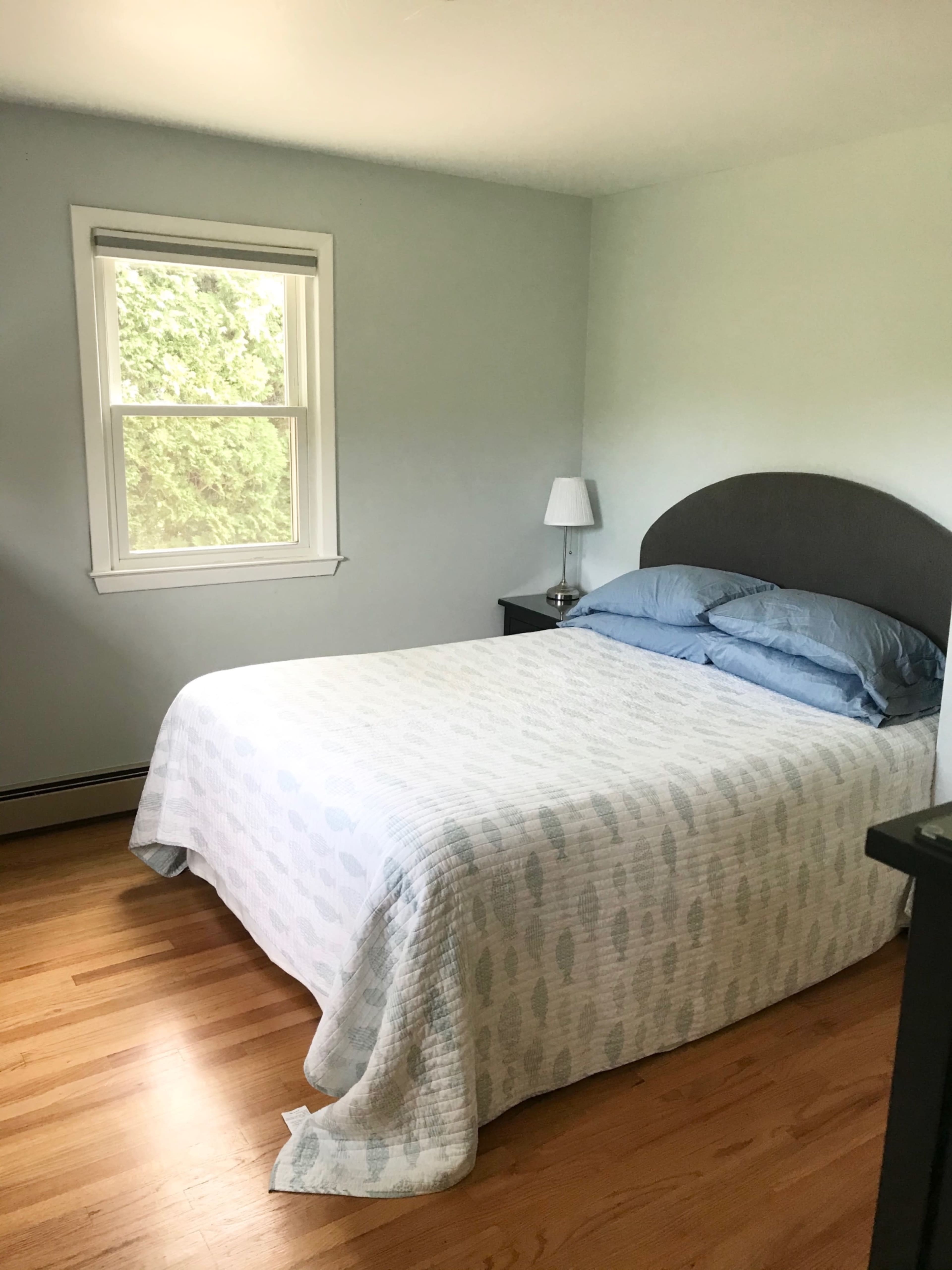 A simple bedroom features a bed with a light quilt, a nightstand with a lamp, and a window allowing natural light.