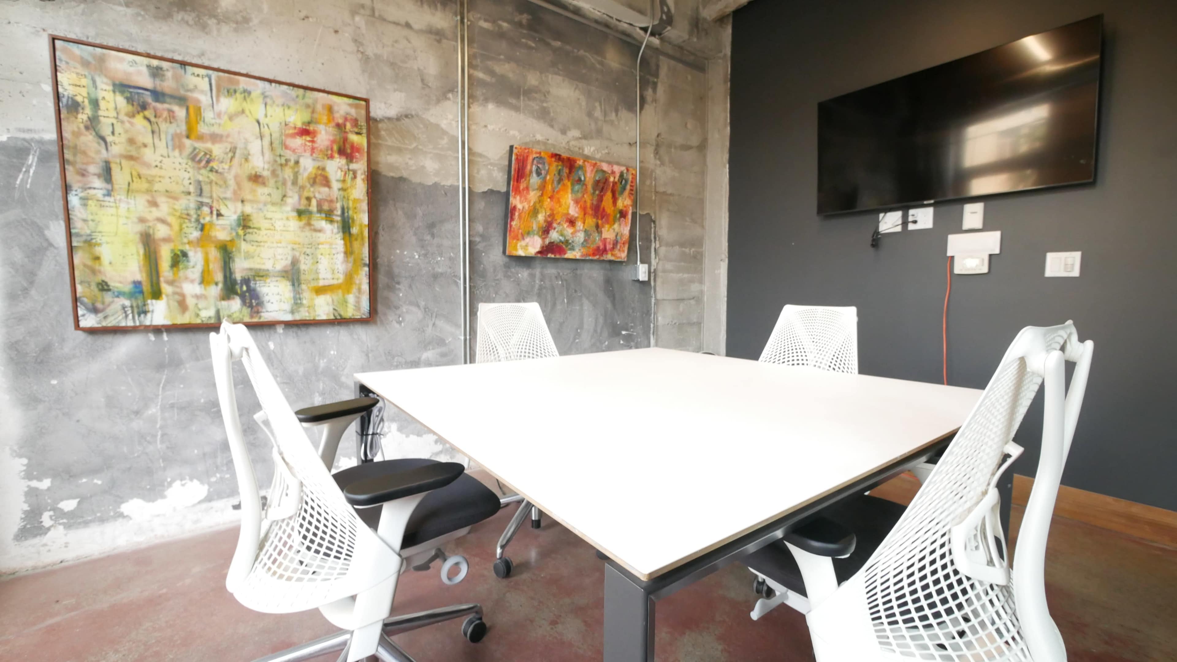 A modern conference room features a large white table surrounded by four ergonomic chairs, with abstract art on the walls and a mounted television.