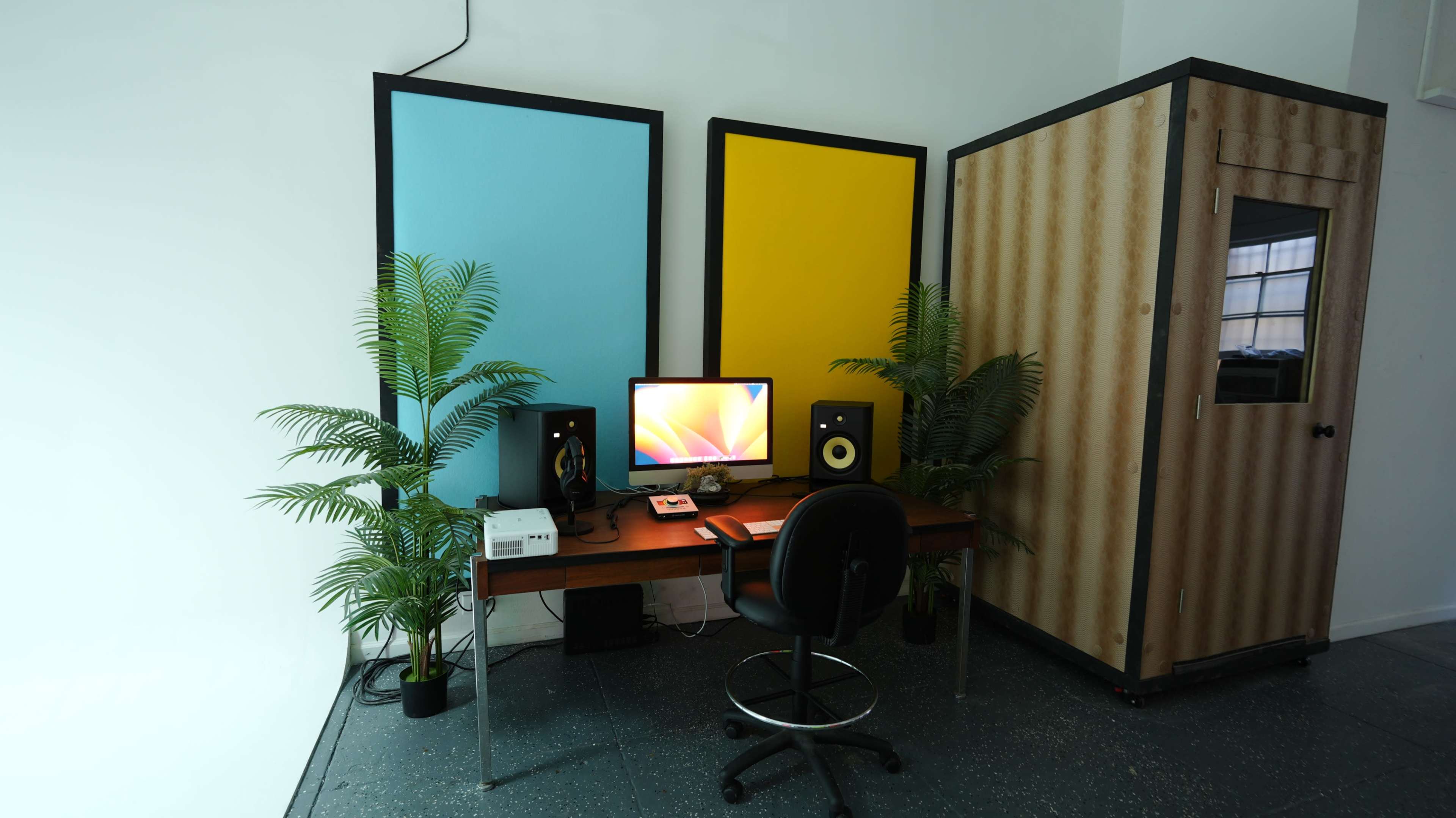 The image shows a small workspace featuring a desk with a computer, speakers, and plants, flanked by two colorful wall panels and a wooden sound booth.