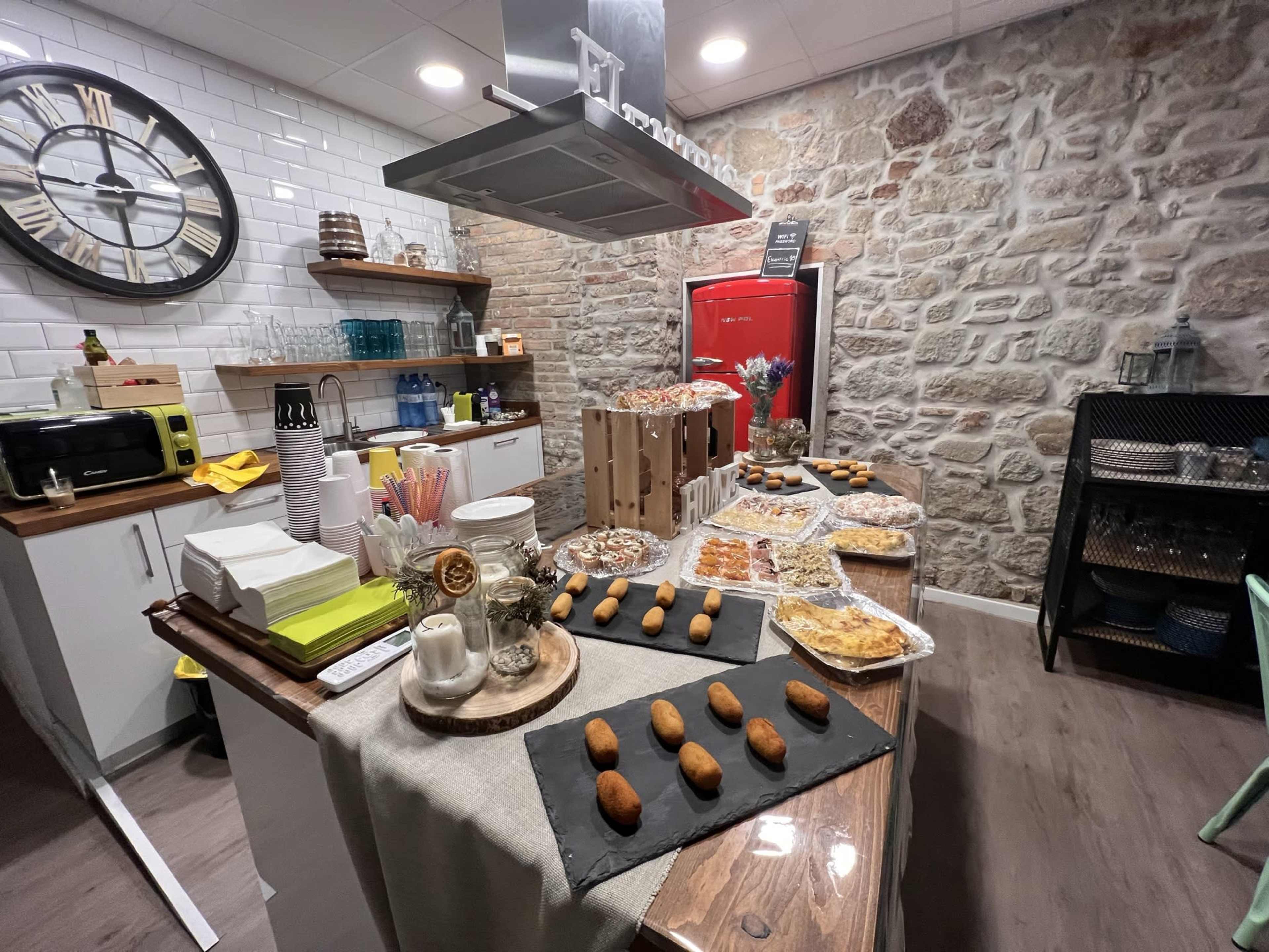 A buffet table filled with various pastries and desserts, set against a rustic stone wall in a well-lit kitchen area.