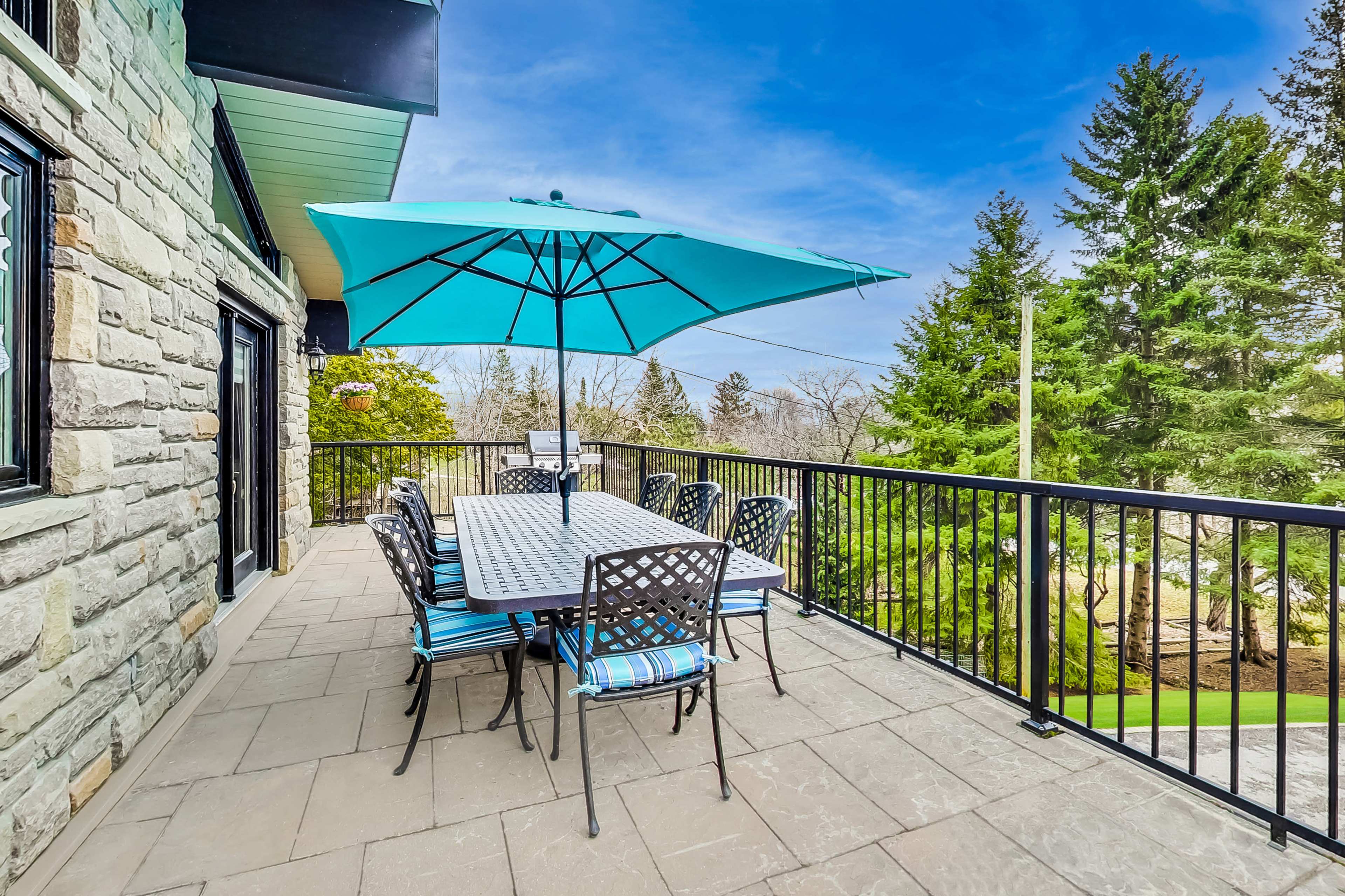 A stone patio features a large table with striped chairs beneath a teal umbrella, overlooking a green landscape and trees.