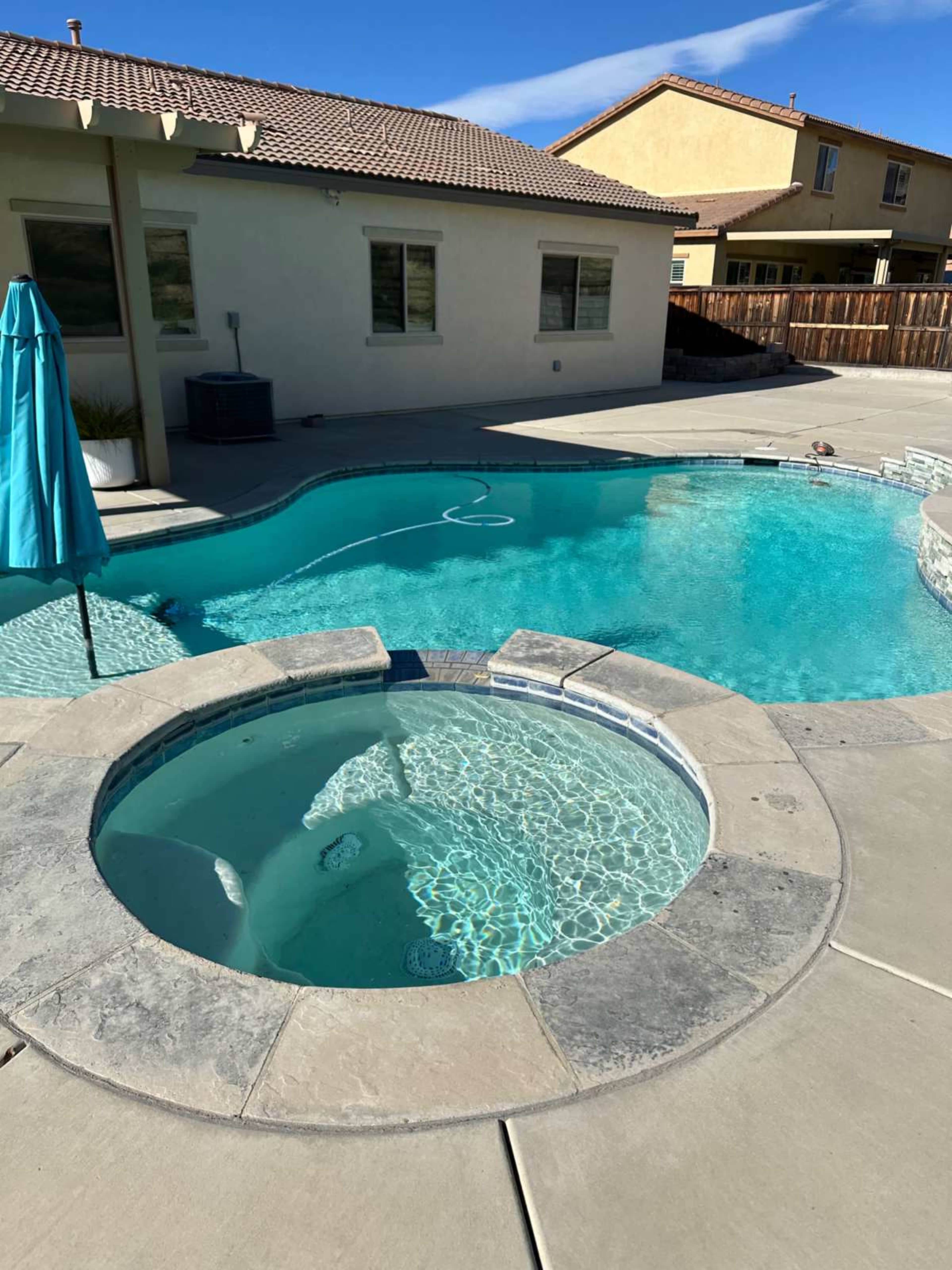The image shows a backyard with a swimming pool and a connected spa, surrounded by a concrete deck and a few residential homes in the background.