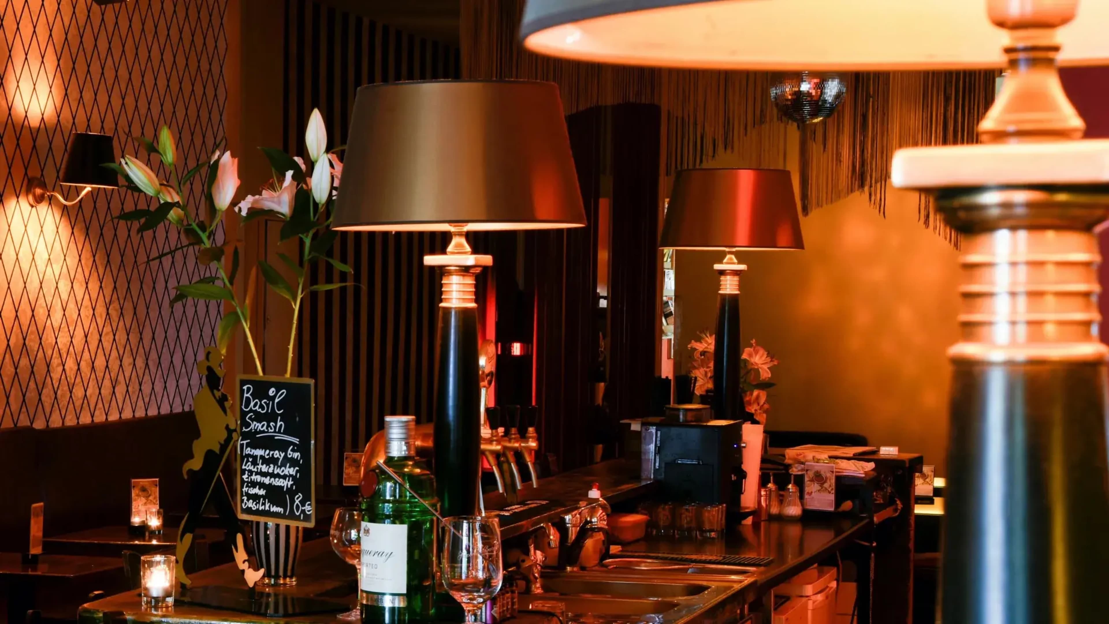 A stylish bar scene features elegant lamps, a chalkboard menu, and a decorative arrangement of plants and bottles.
