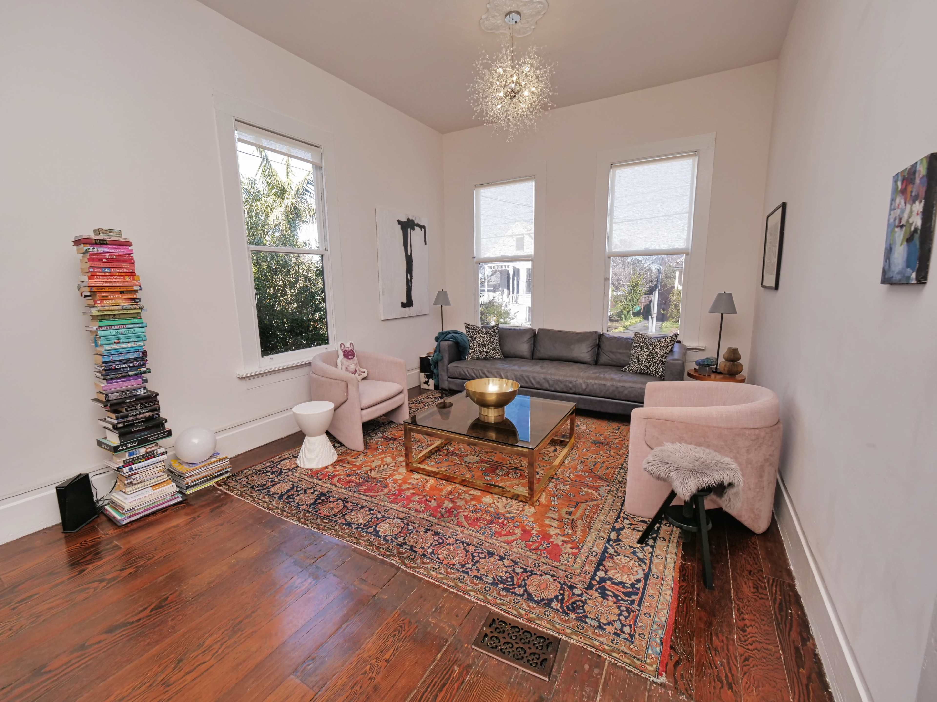 The image shows a living room with a gray sofa, two light pink chairs, a glass coffee table, and a tall stack of books next to a window.