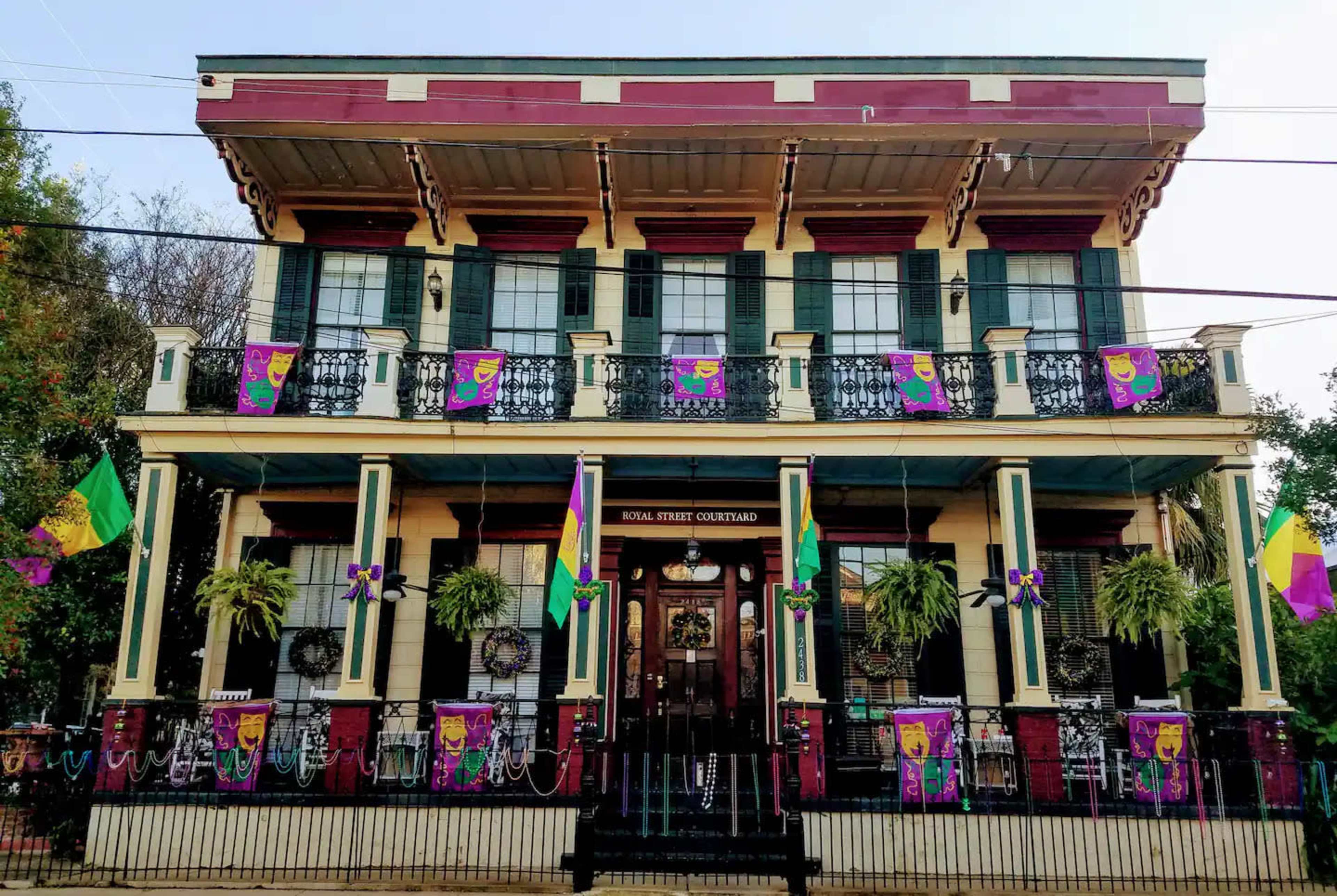 The image shows a two-story building adorned with colorful Mardi Gras decorations, including banners and wreaths, set within a wrought iron fence.