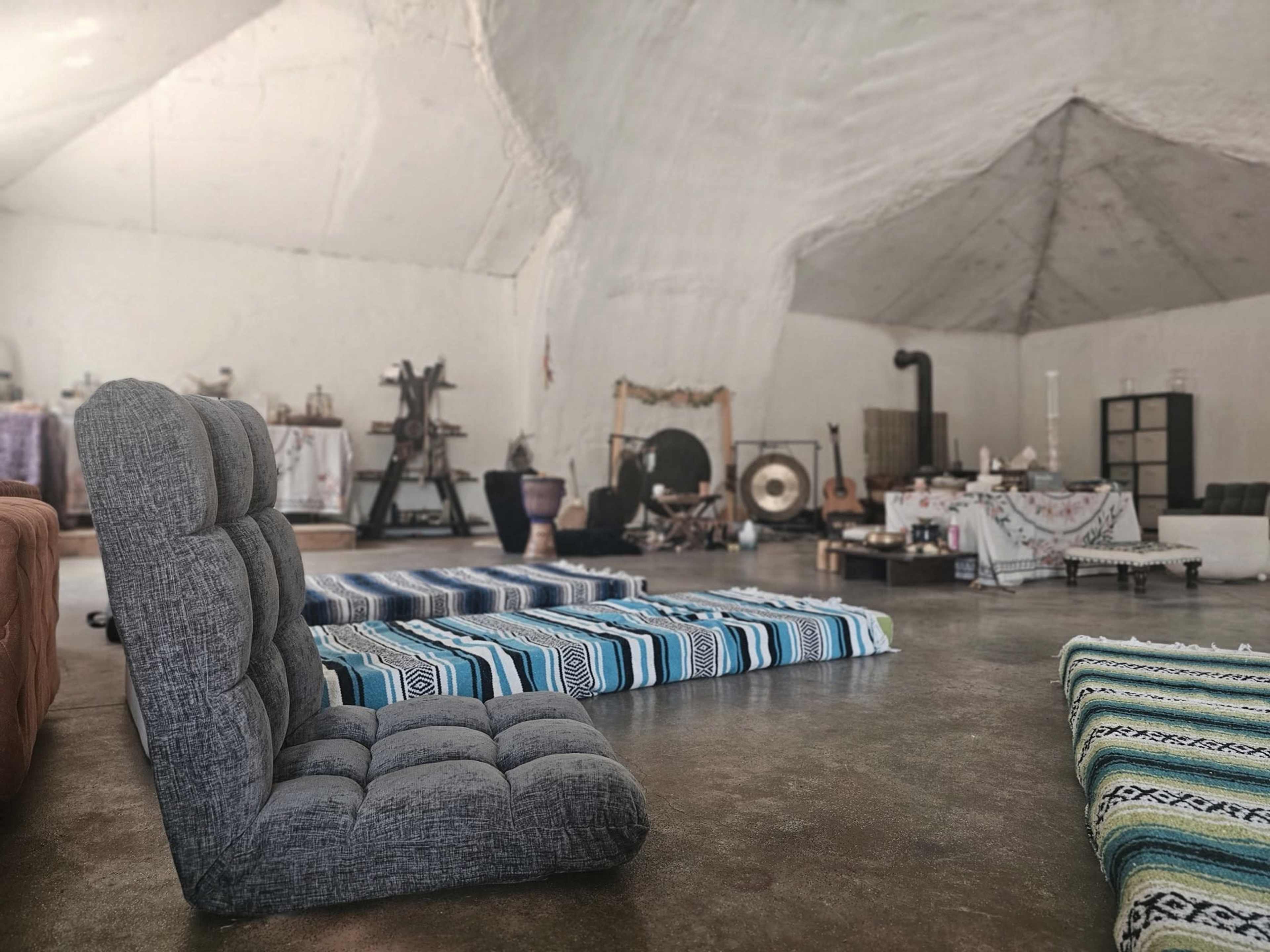 The image shows a spacious, minimalist room with several floor cushions arranged on a concrete floor, under a vaulted ceiling, and with various instruments and decor items scattered around.