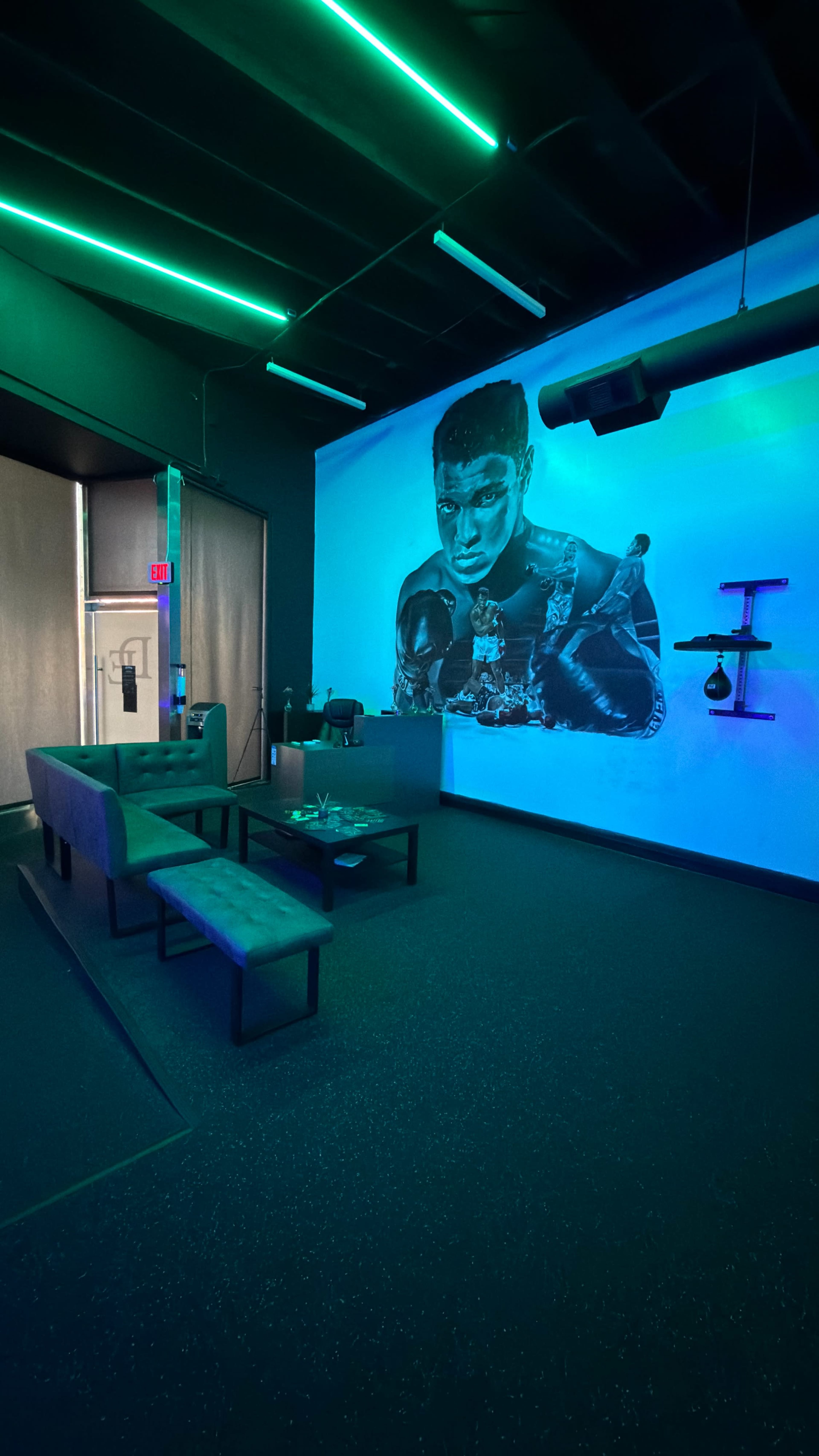The image shows a dimly lit room with a large mural of a boxer on the wall, featuring modern seating and ambient neon lighting.