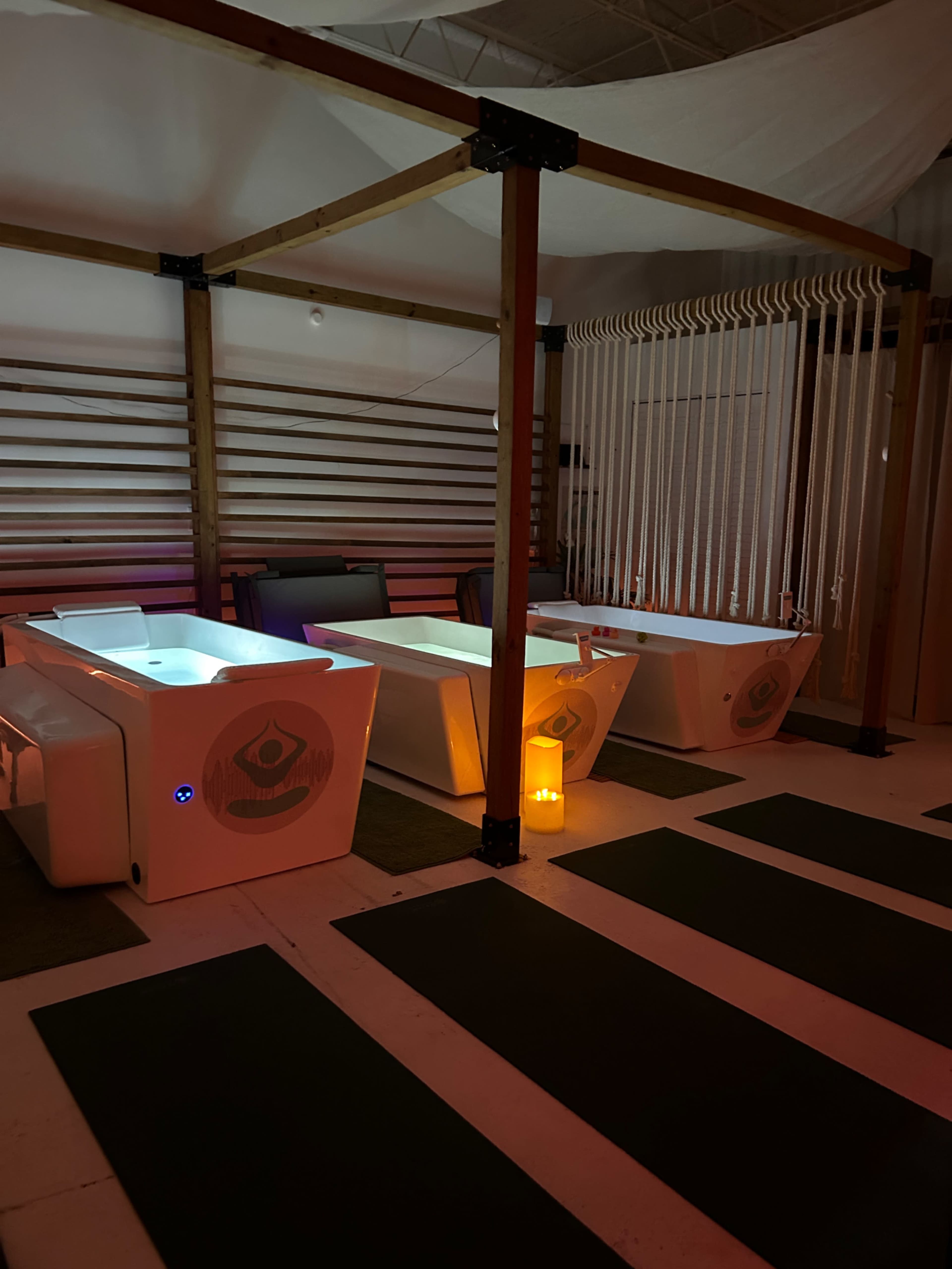 The image shows a serene spa setting with three illuminated therapeutic tubs and yoga mats arranged on the floor.