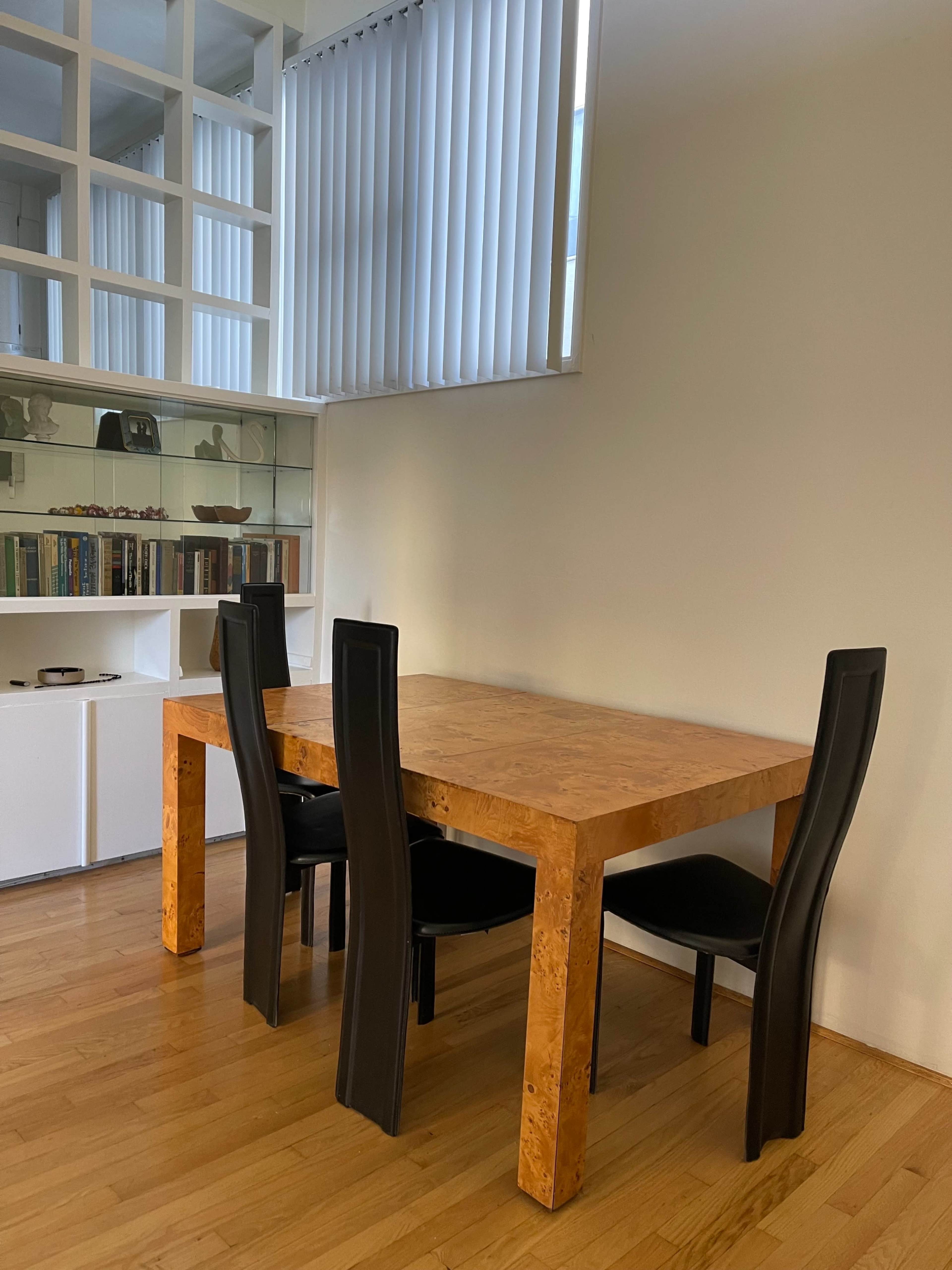A wooden table with a textured surface is surrounded by four black chairs in a minimalist room with white walls and large vertical blinds.