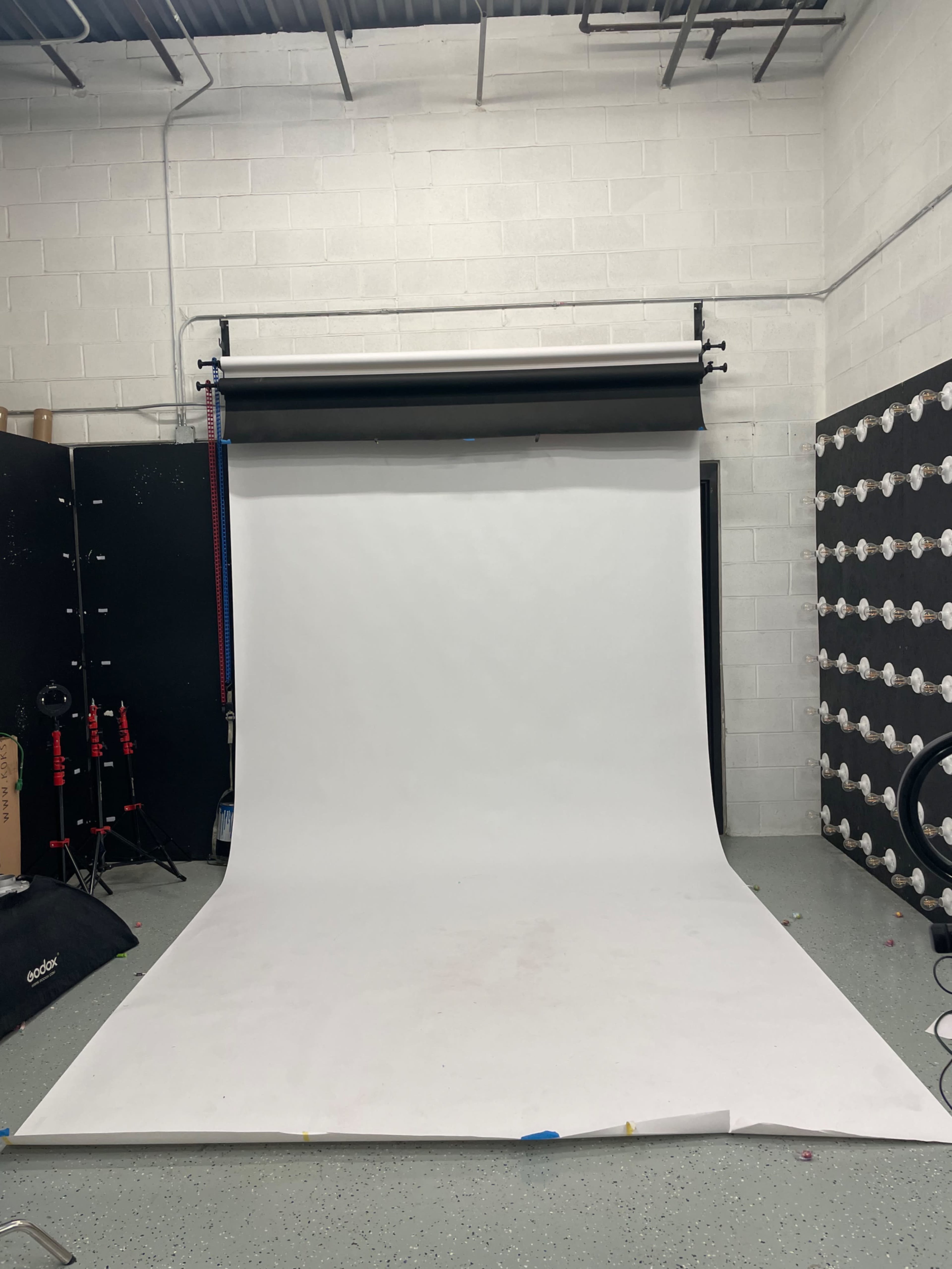 The image shows a photography studio with a white backdrop set up against a textured wall, and various equipment arranged in the background.