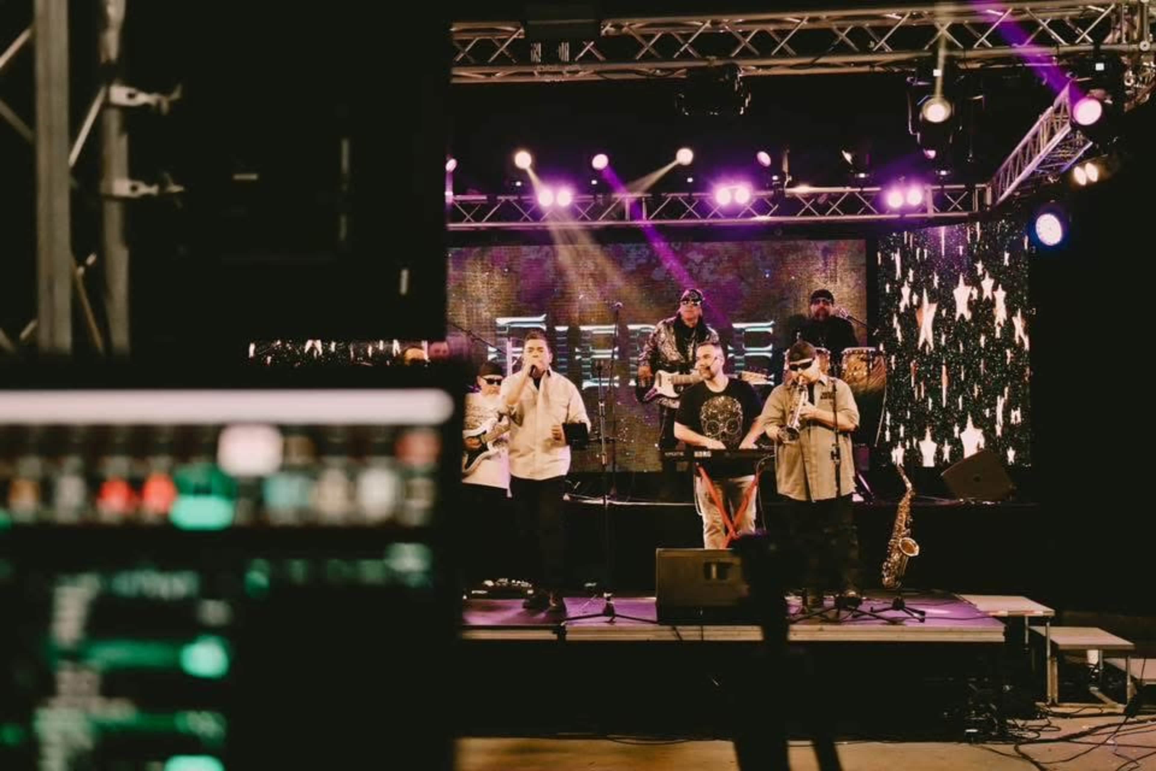 A music group performs on stage with various instruments while a sound mixing console is visible in the foreground.