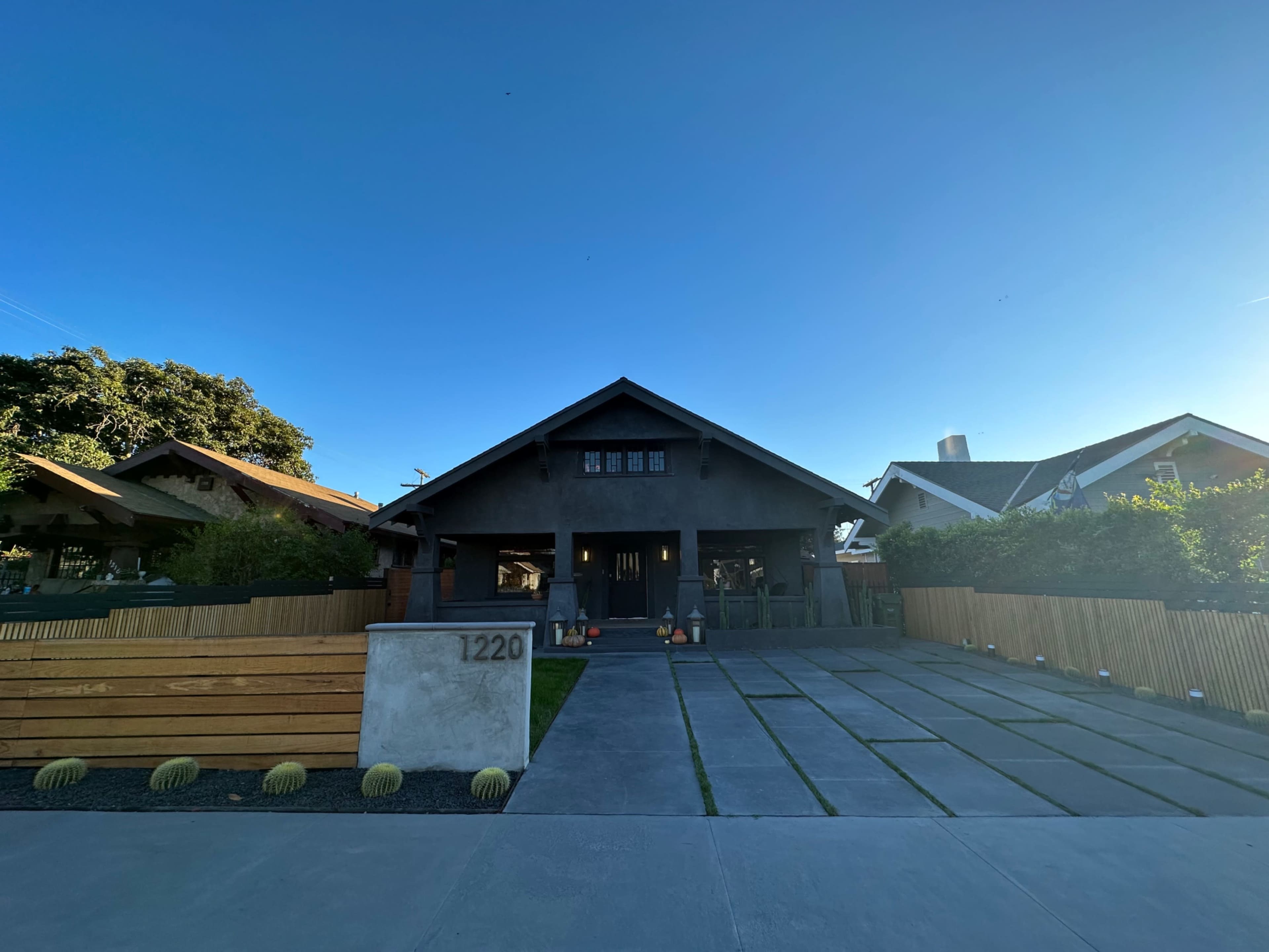The image shows a modern house with a dark exterior and a landscaped front yard featuring concrete paving and wooden fencing.