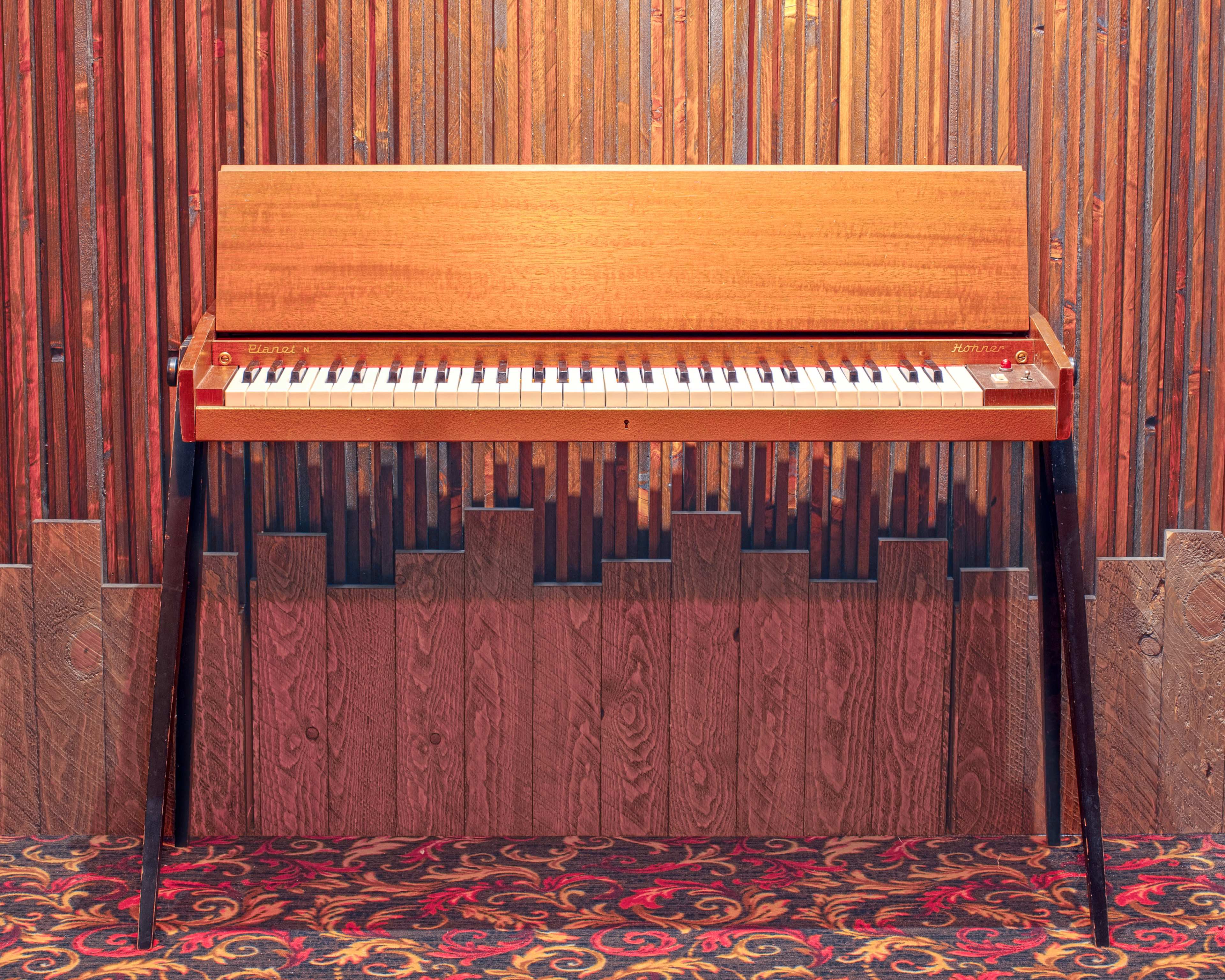A wooden electric piano stands against a textured wood-paneled wall with a patterned carpet beneath it.