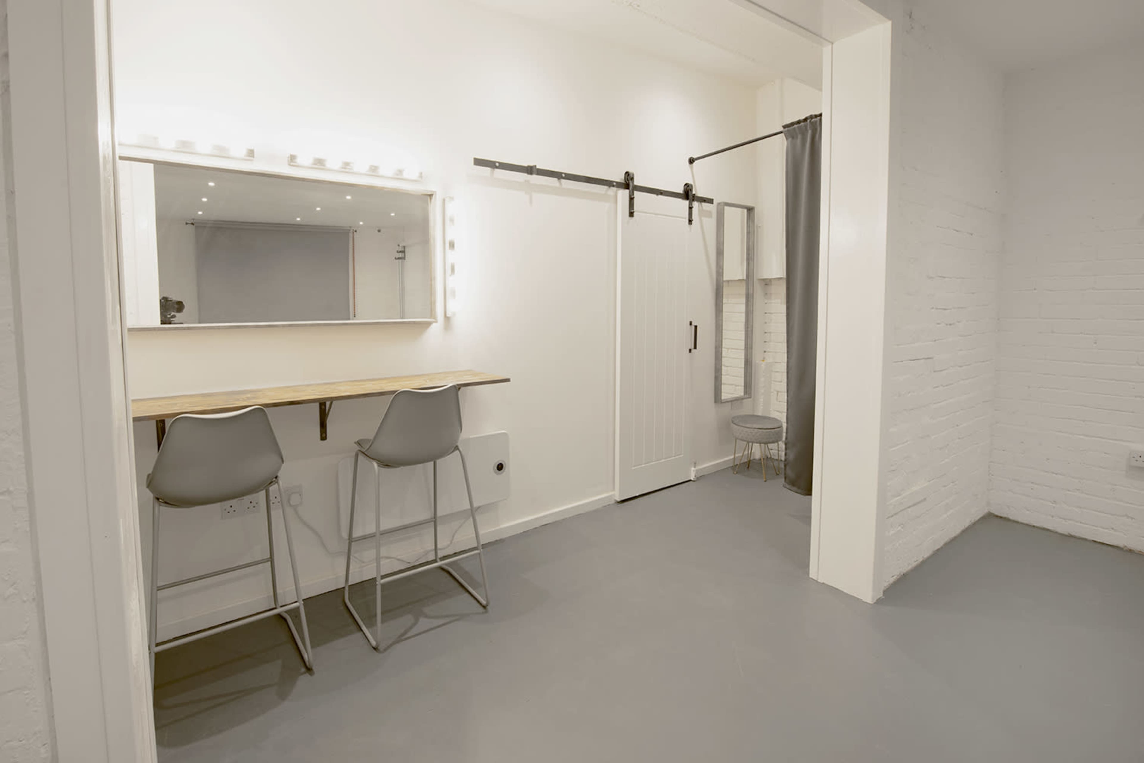 A well-lit, minimalist room with a mirror and two tall chairs next to a vanity, a sliding barn door, and a curtain.