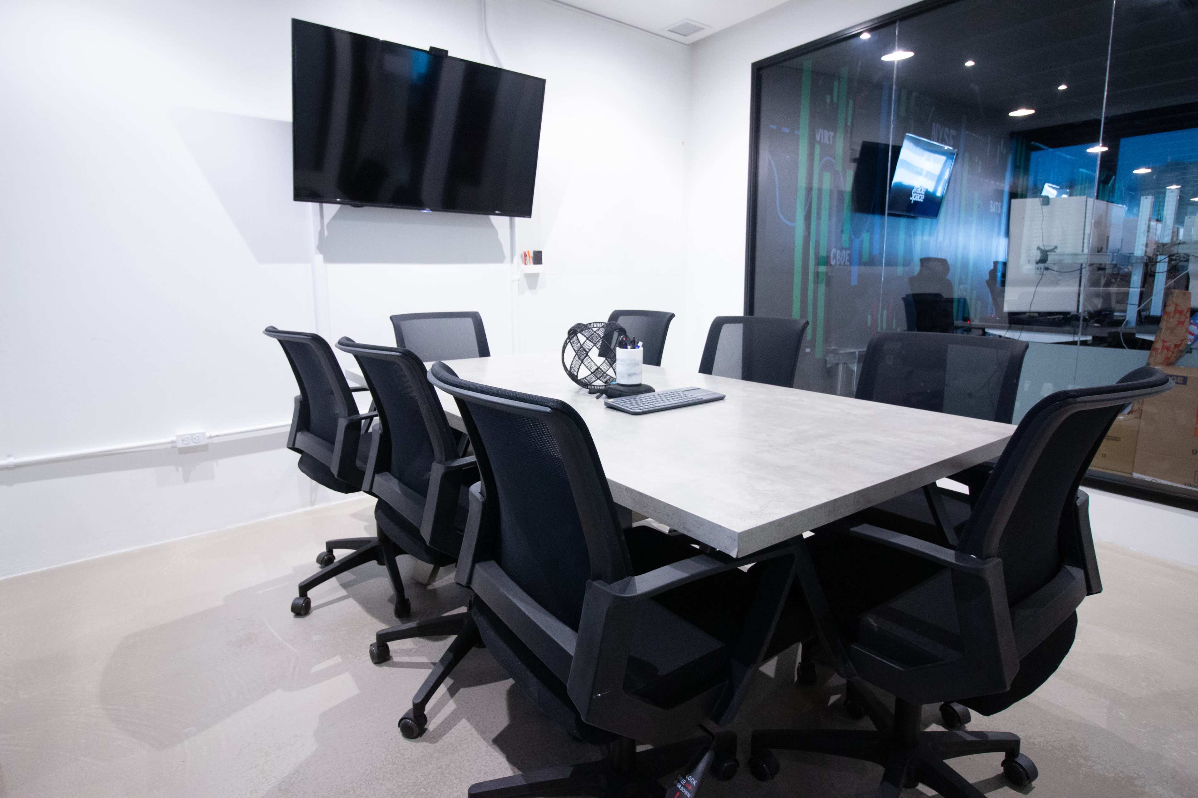 A modern conference room features a large gray table surrounded by six ergonomic chairs and a wall-mounted television.