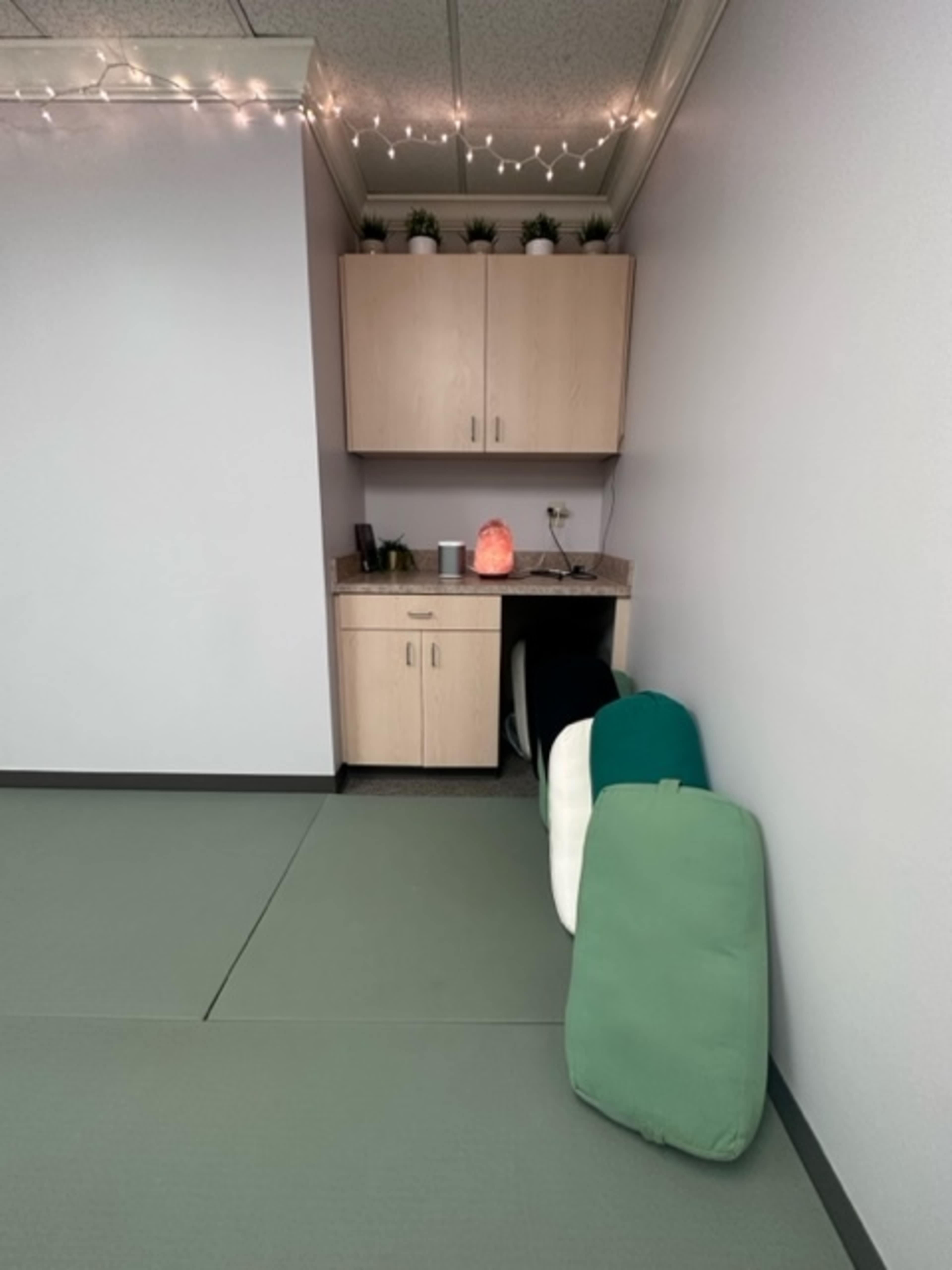 A corner of a room with a countertop, cabinets, and a few cushions stacked against the wall, alongside a salt lamp and some potted plants.