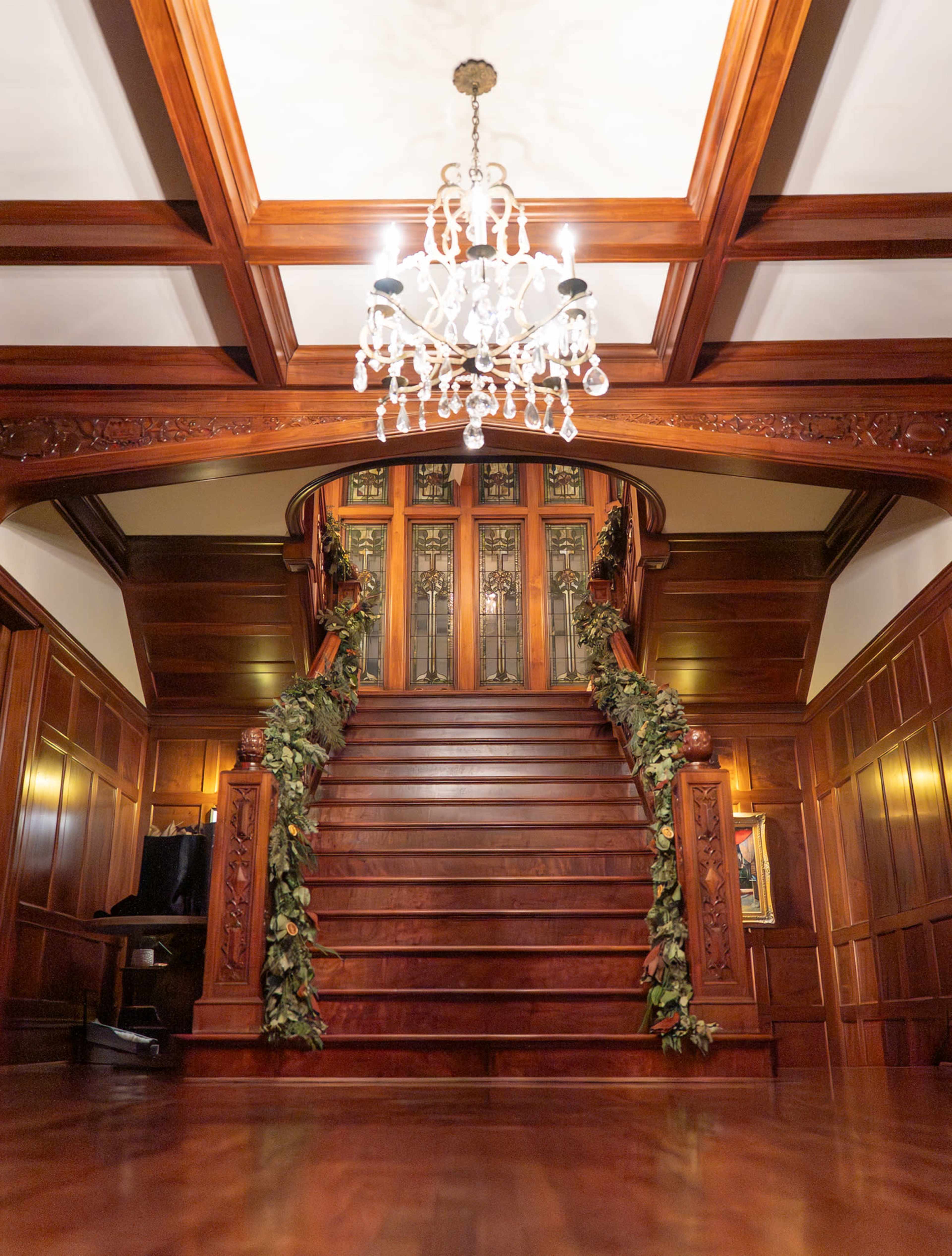 The image shows a wooden staircase with ornate details, leading to an upper landing, illuminated by a crystal chandelier above.