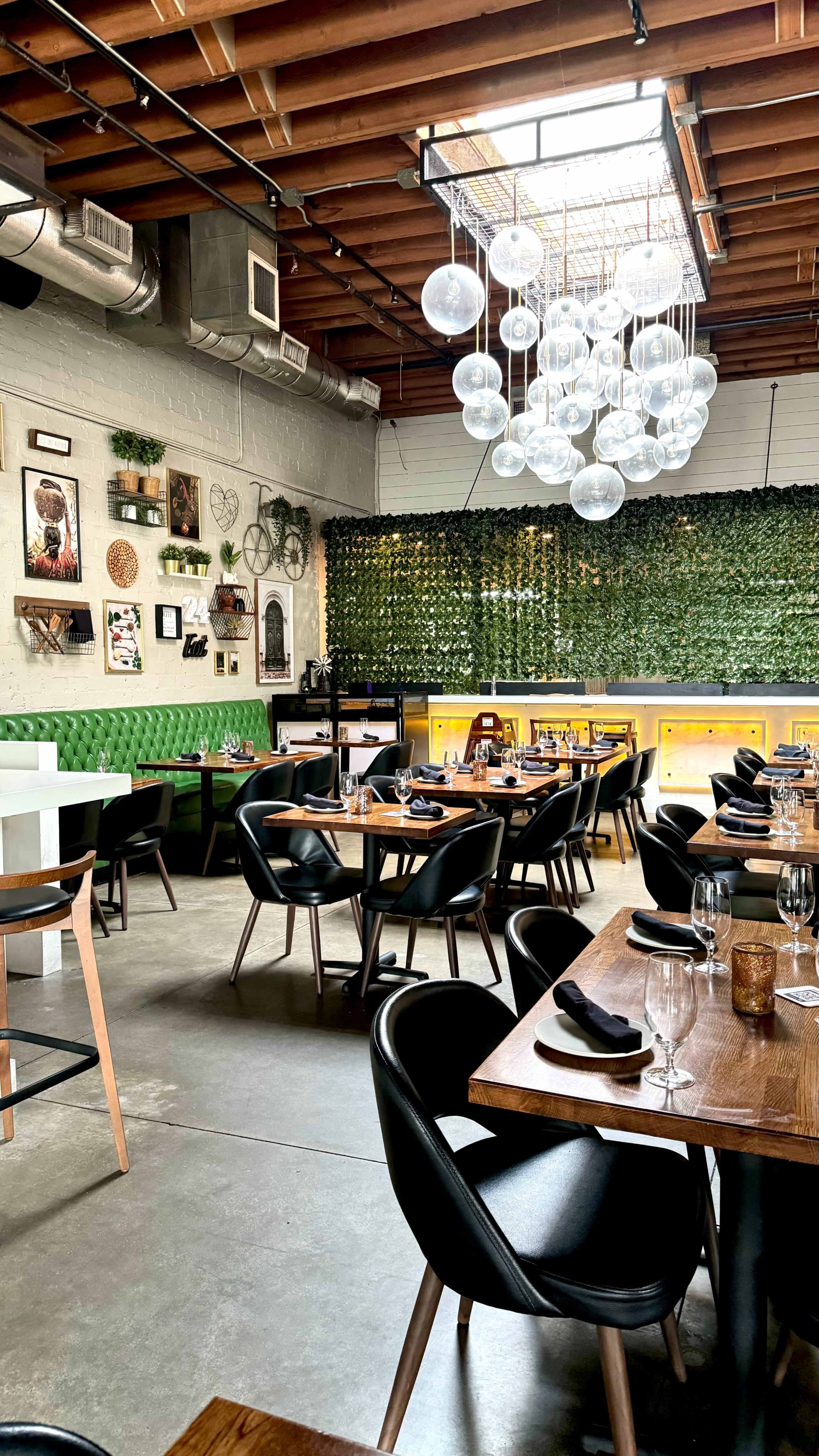 The image shows a modern restaurant interior with wooden tables, black chairs, green decor on the walls, and a cluster of decorative light fixtures hanging from the ceiling.