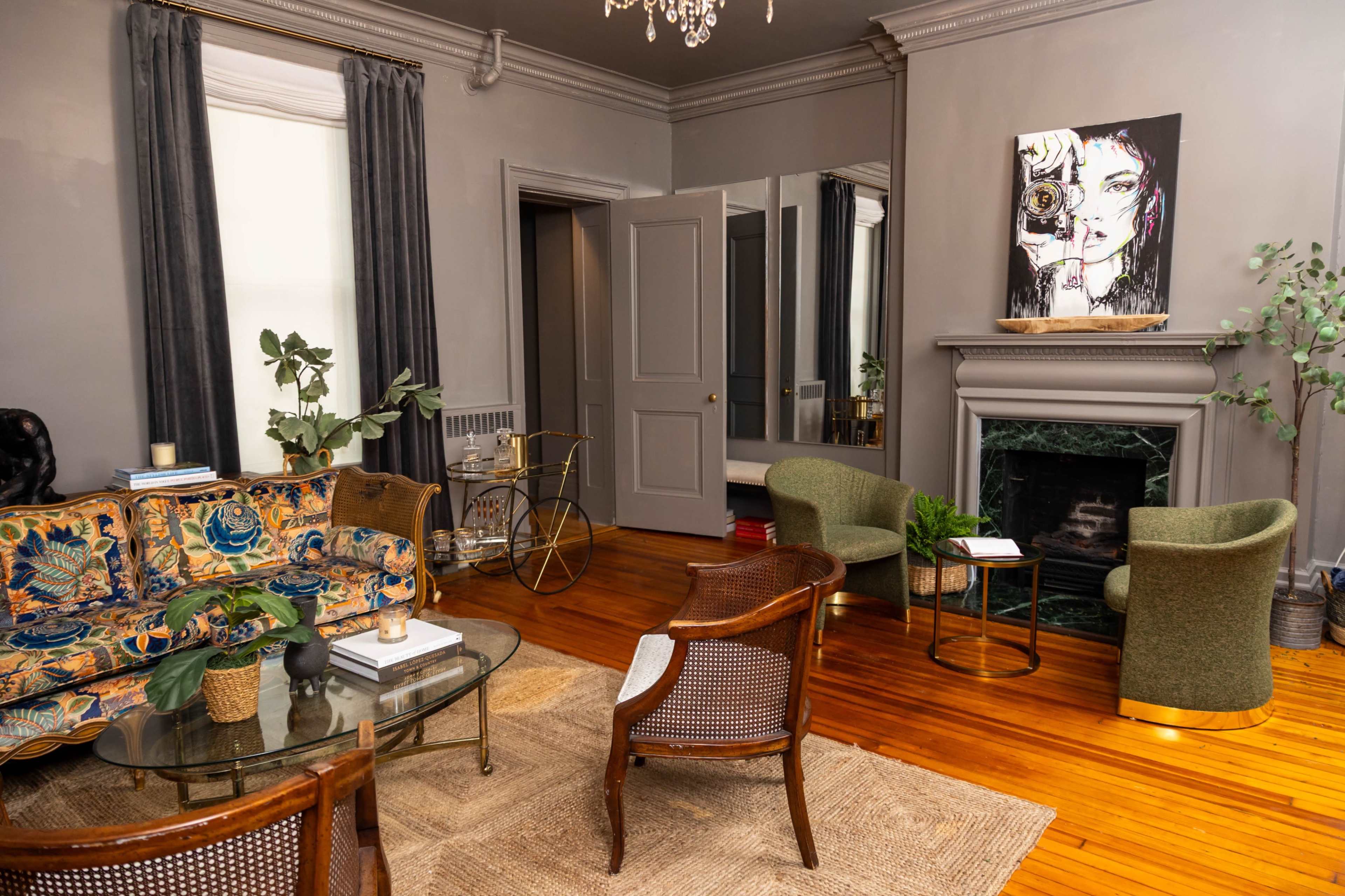 The image shows a stylish living room with a mix of vintage and modern furniture, including a colorful sofa, armchairs, and a glass coffee table, set against a backdrop of gray walls and a fireplace.