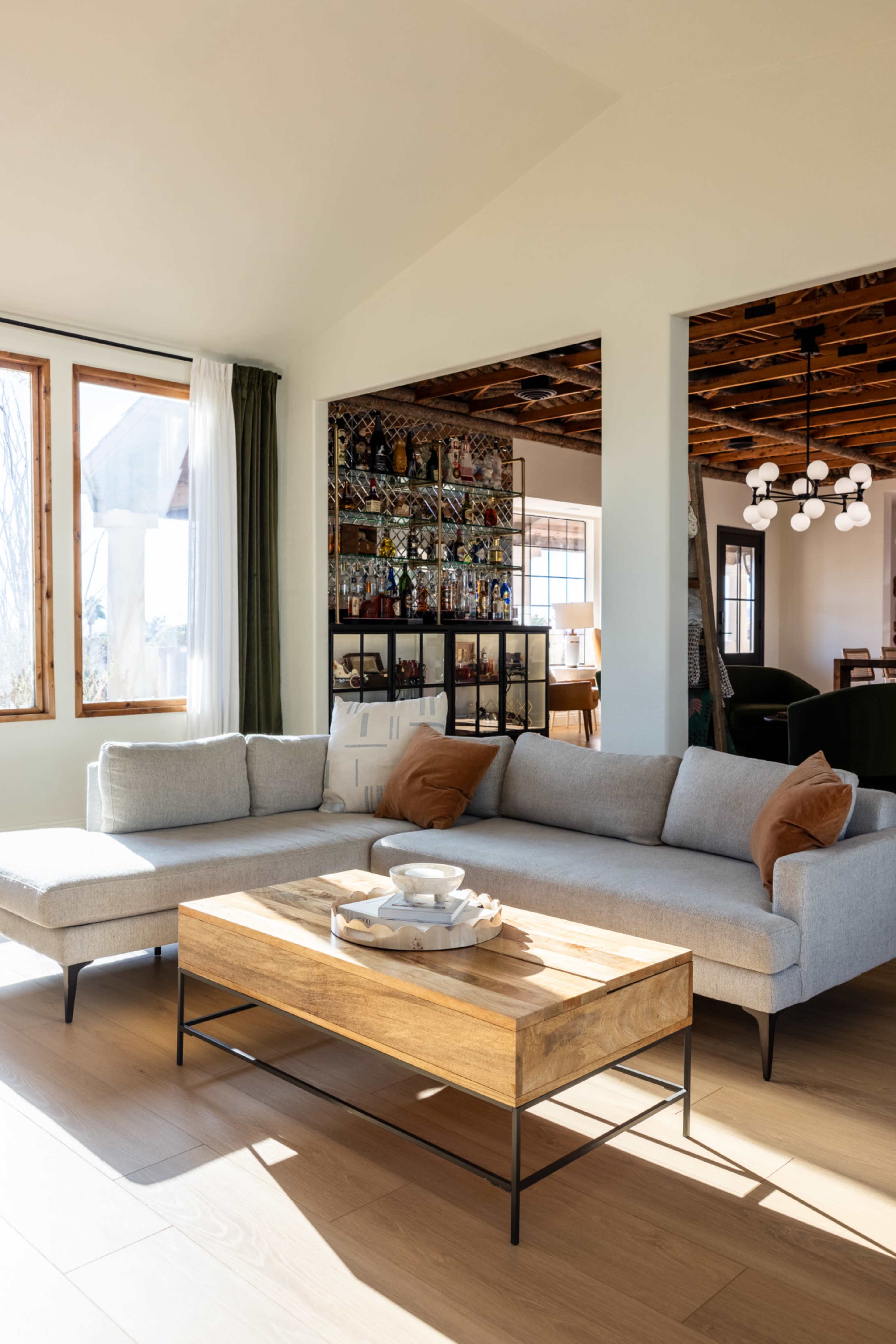 The image shows a modern living room featuring a gray sectional sofa, a wooden coffee table, and large windows that allow natural light to fill the space.