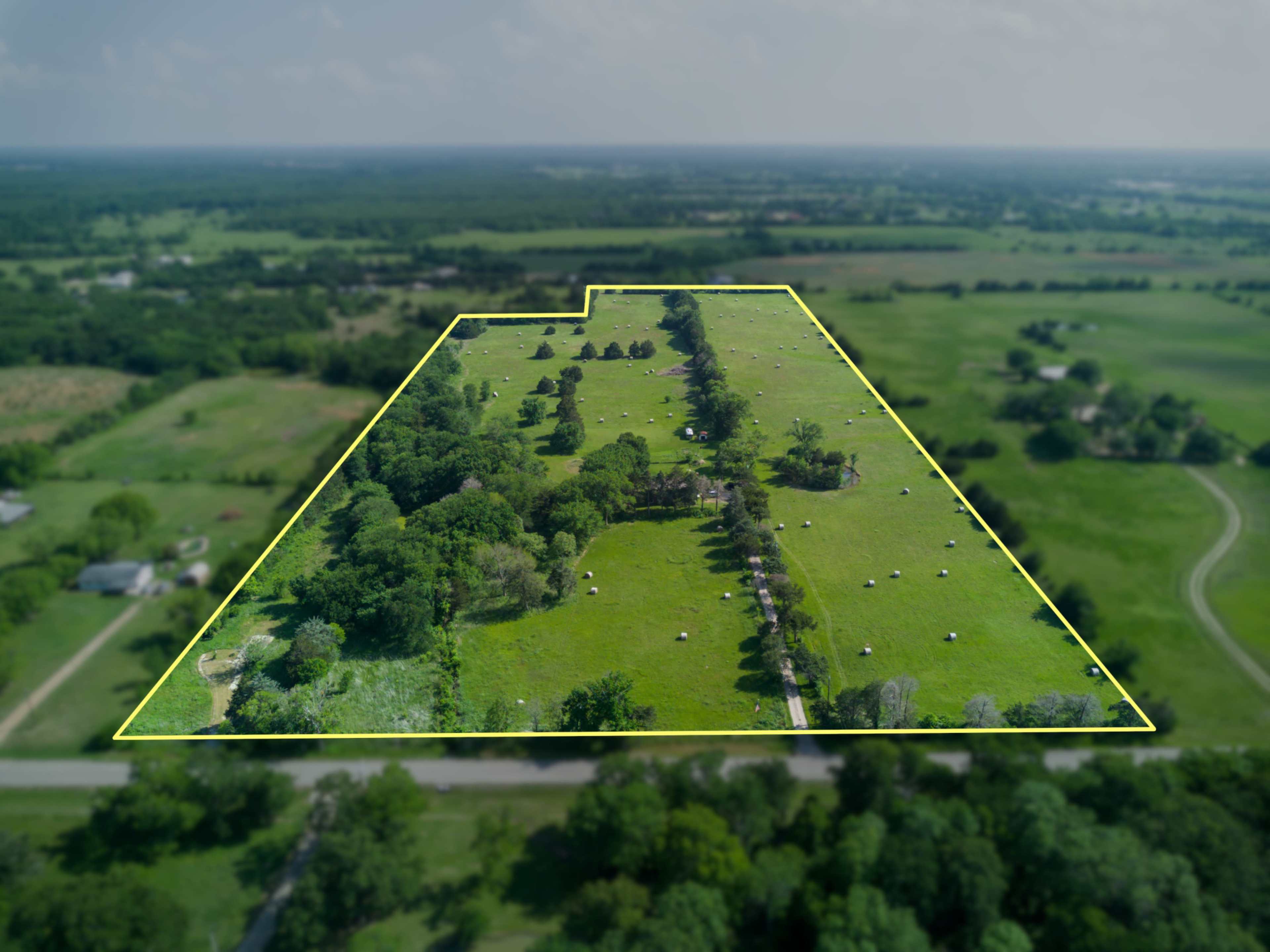 The image shows an aerial view of a large, rectangular plot of green land bordered by a yellow outline, featuring scattered trees and hay bales.