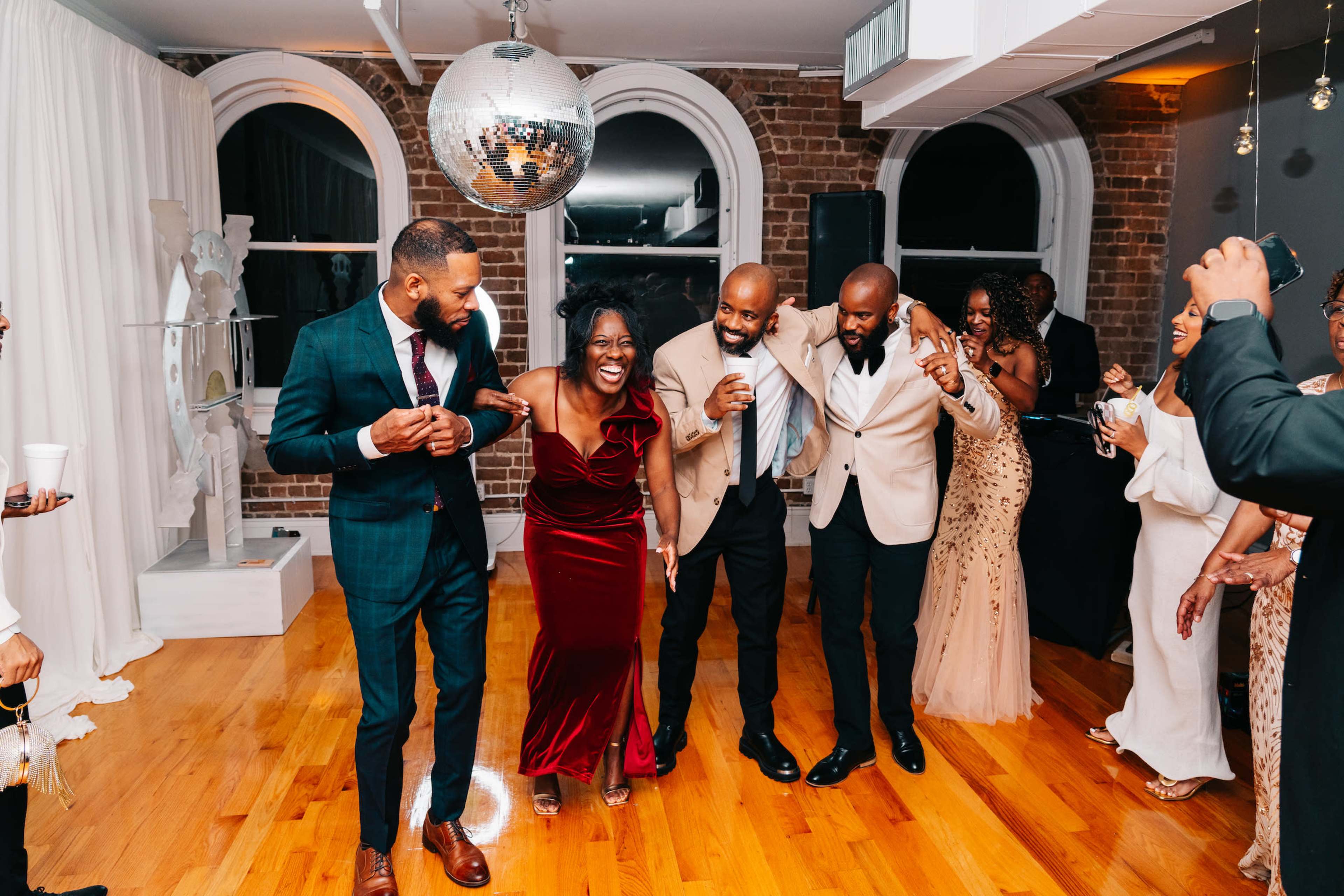 A group of five people dances joyfully in a well-lit room with wooden floors and a disco ball overhead.