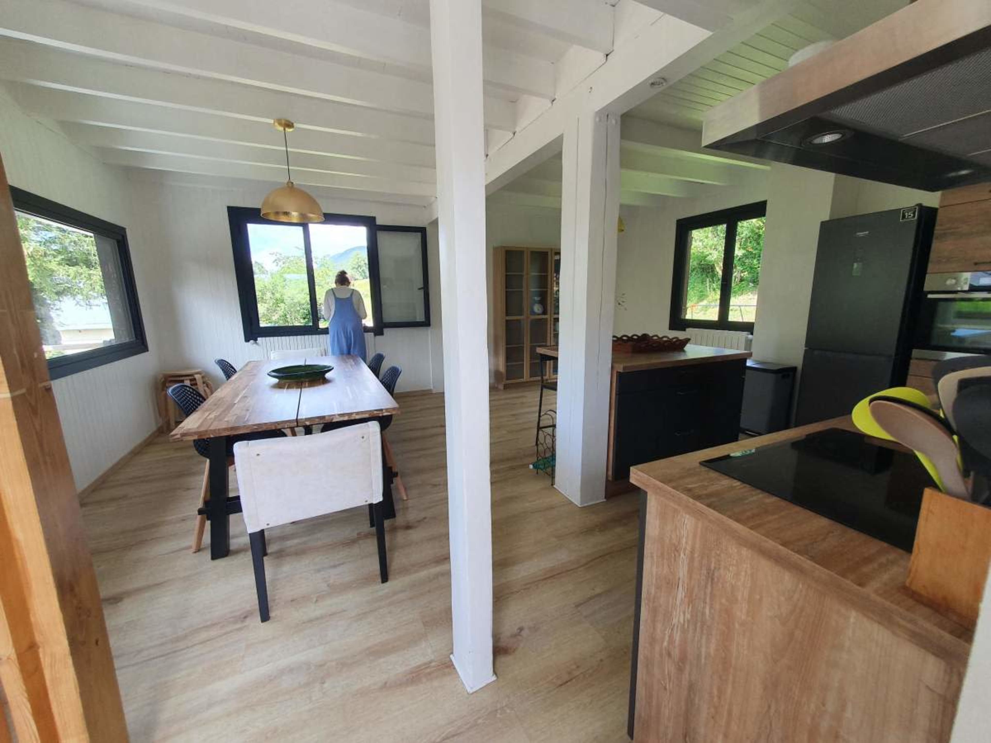 The image shows a modern dining area with a wooden table and chairs, a kitchen counter, and large windows that let in natural light.