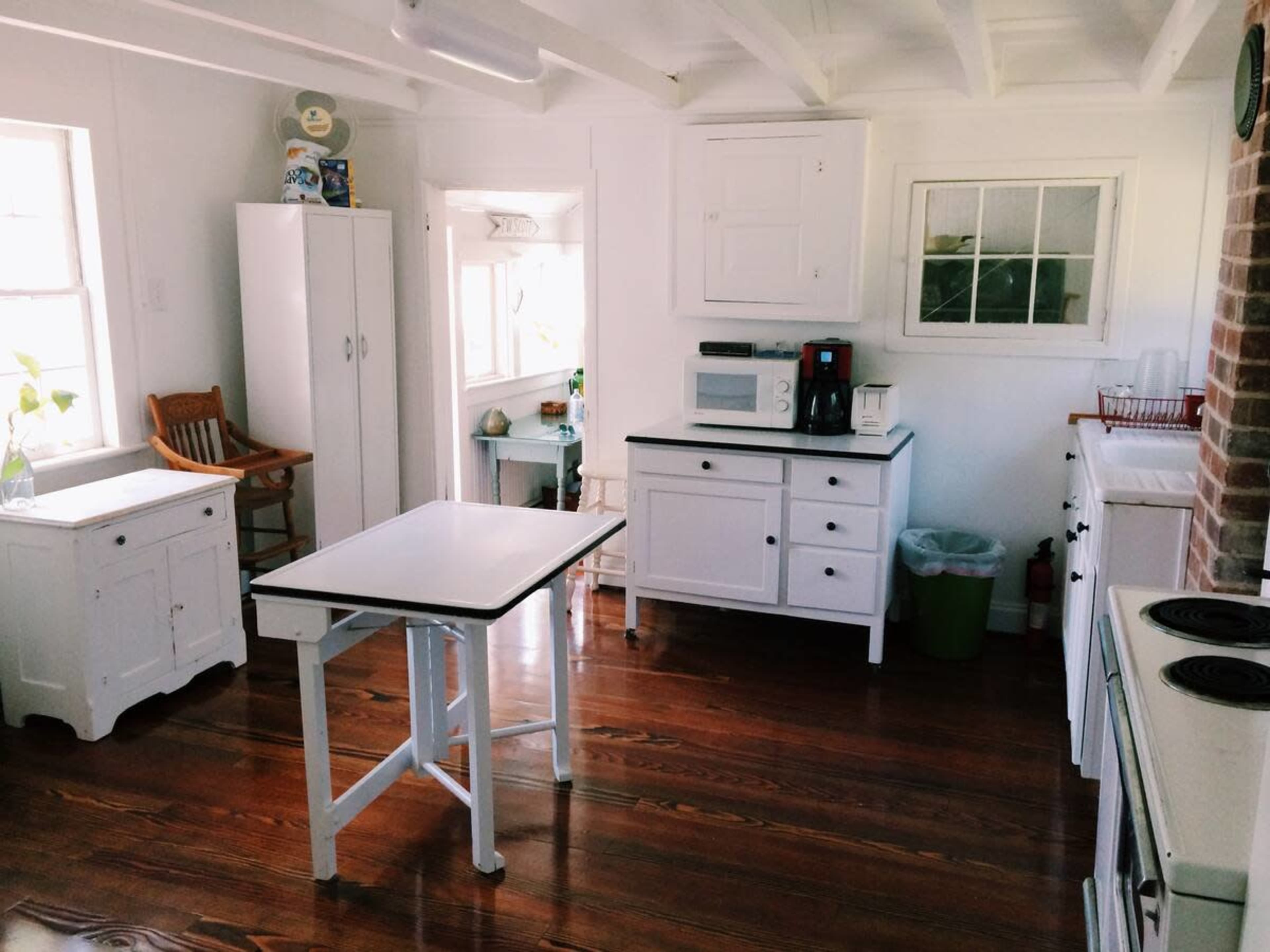 A bright kitchen with white cabinets, a table, and a coffee maker, featuring wooden floors and a window that lets in natural light.