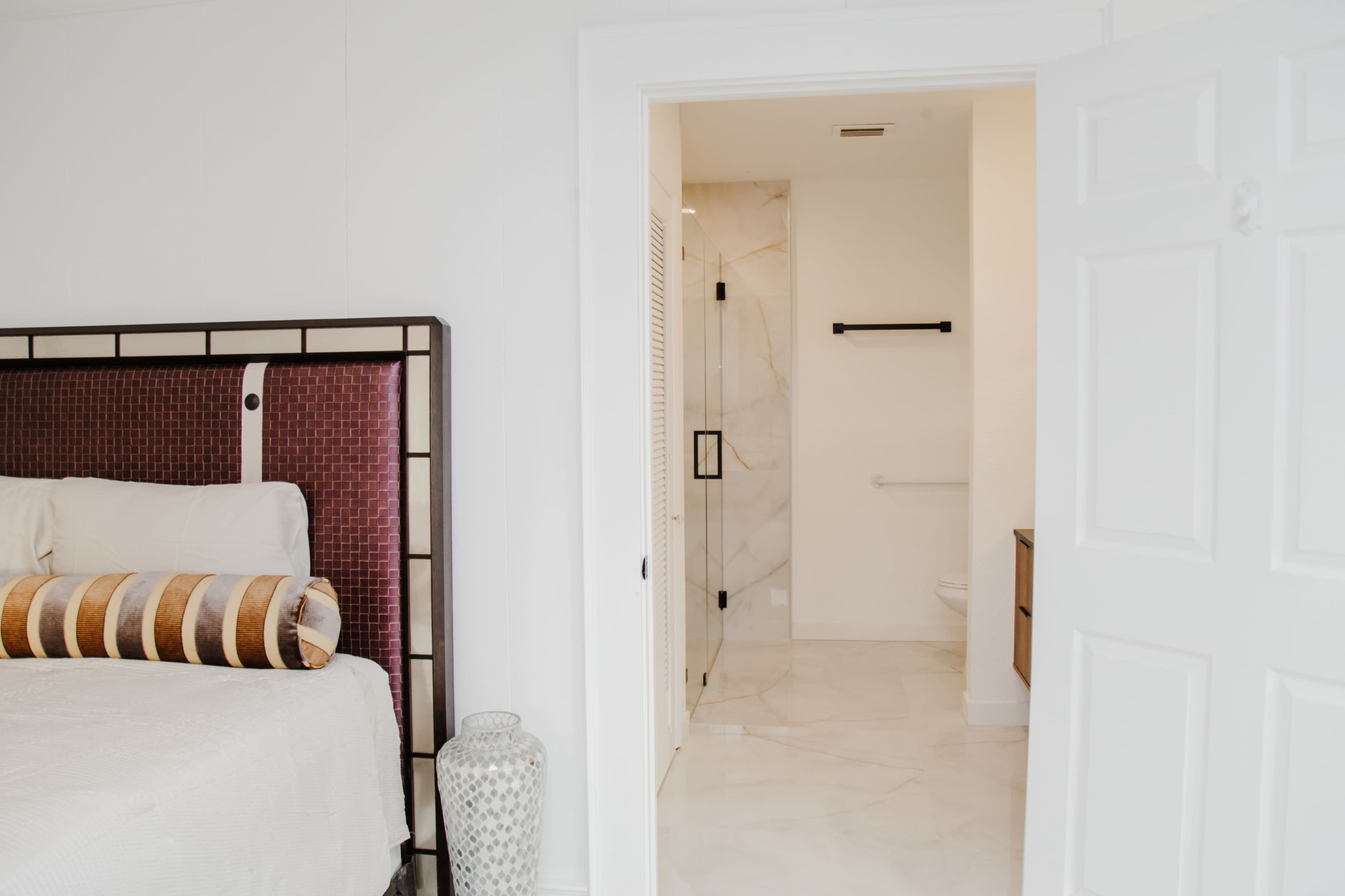 A modern bedroom features a large bed with a maroon headboard and a visible bathroom beyond an open door.