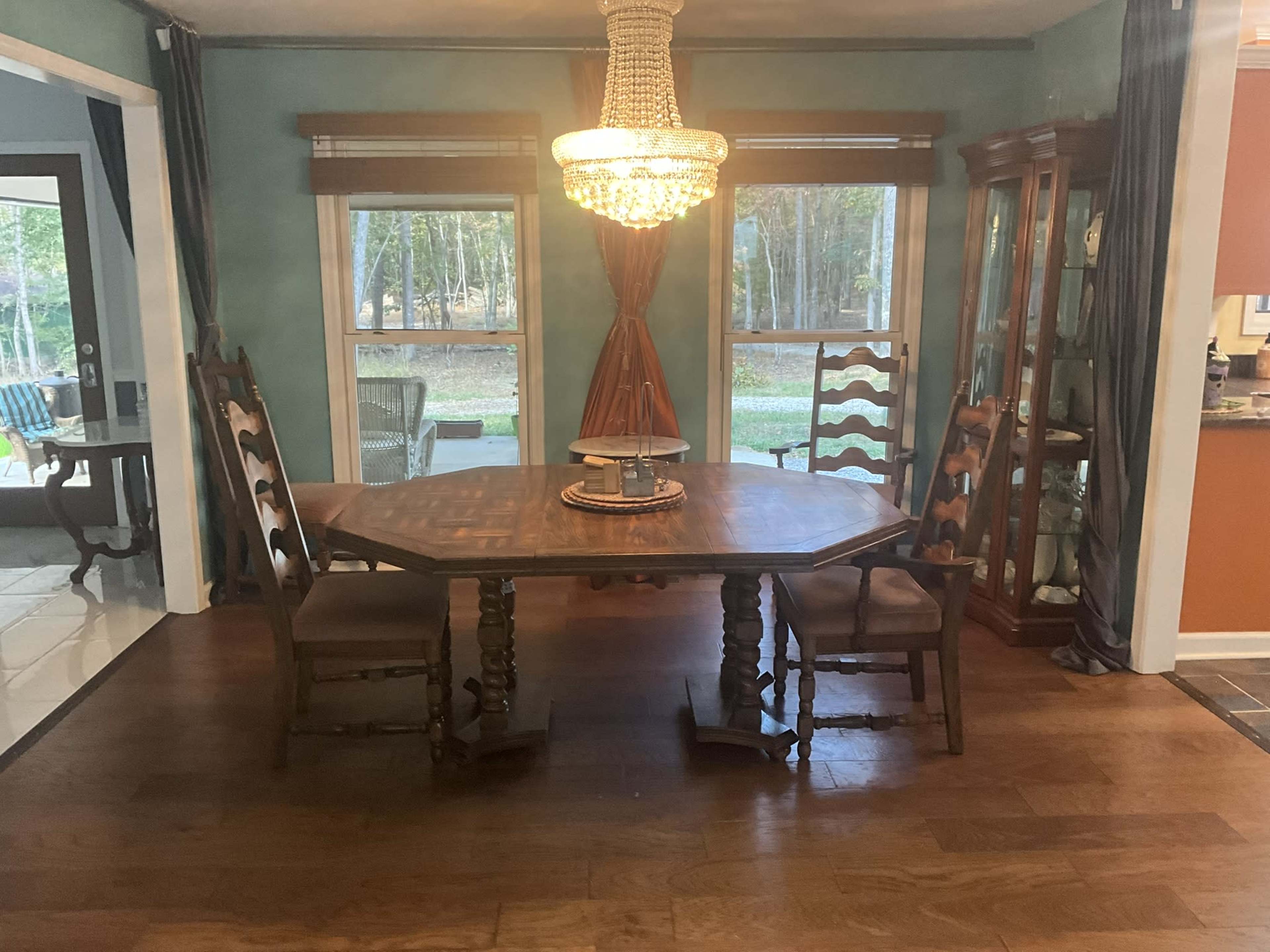 The image shows a wooden dining table with four chairs under a chandelier in a room with large windows overlooking a wooded area.