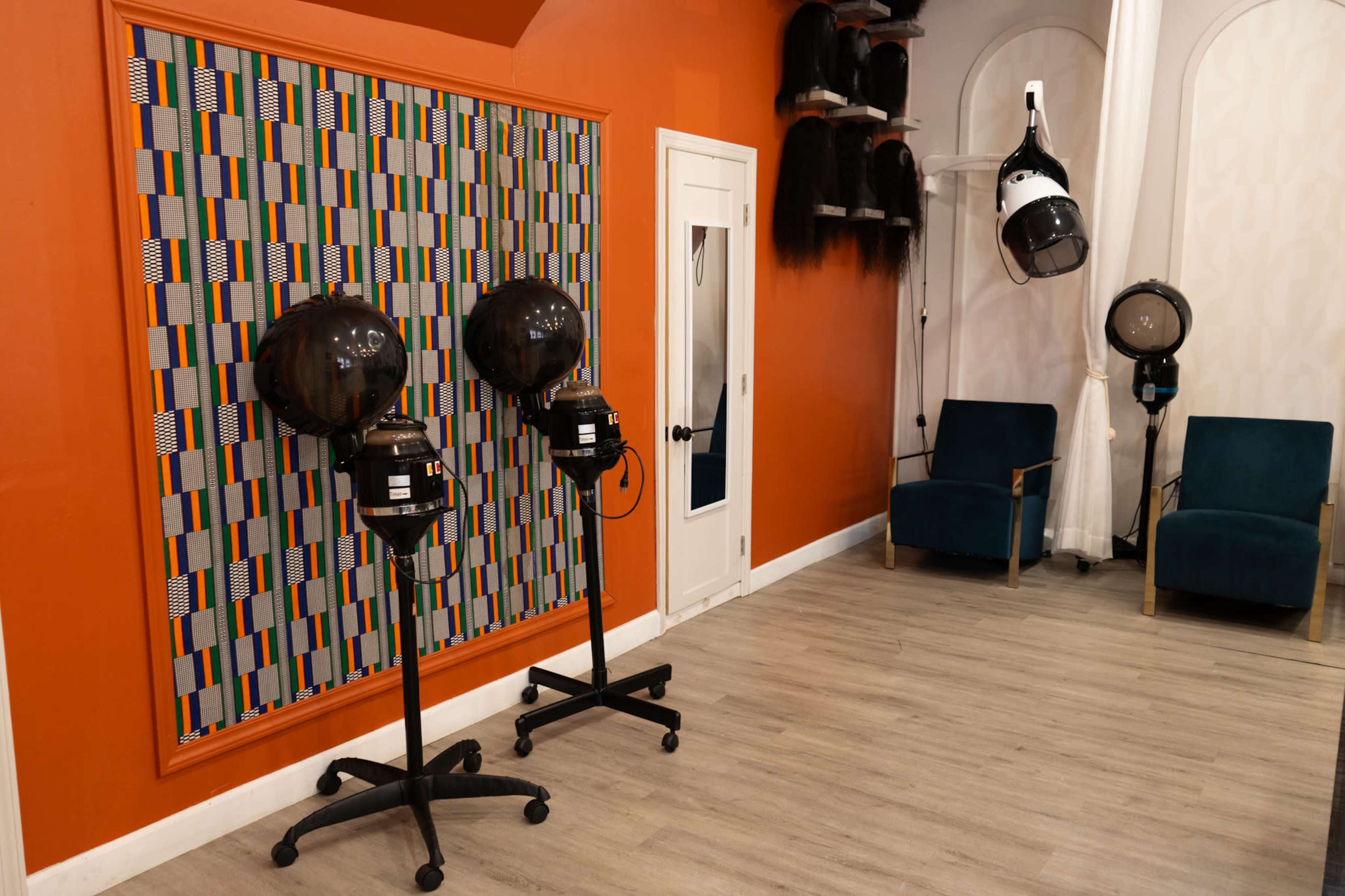The image shows a hair salon interior featuring two hair dryers on stands, a patterned wall, and two blue chairs against an orange wall.
