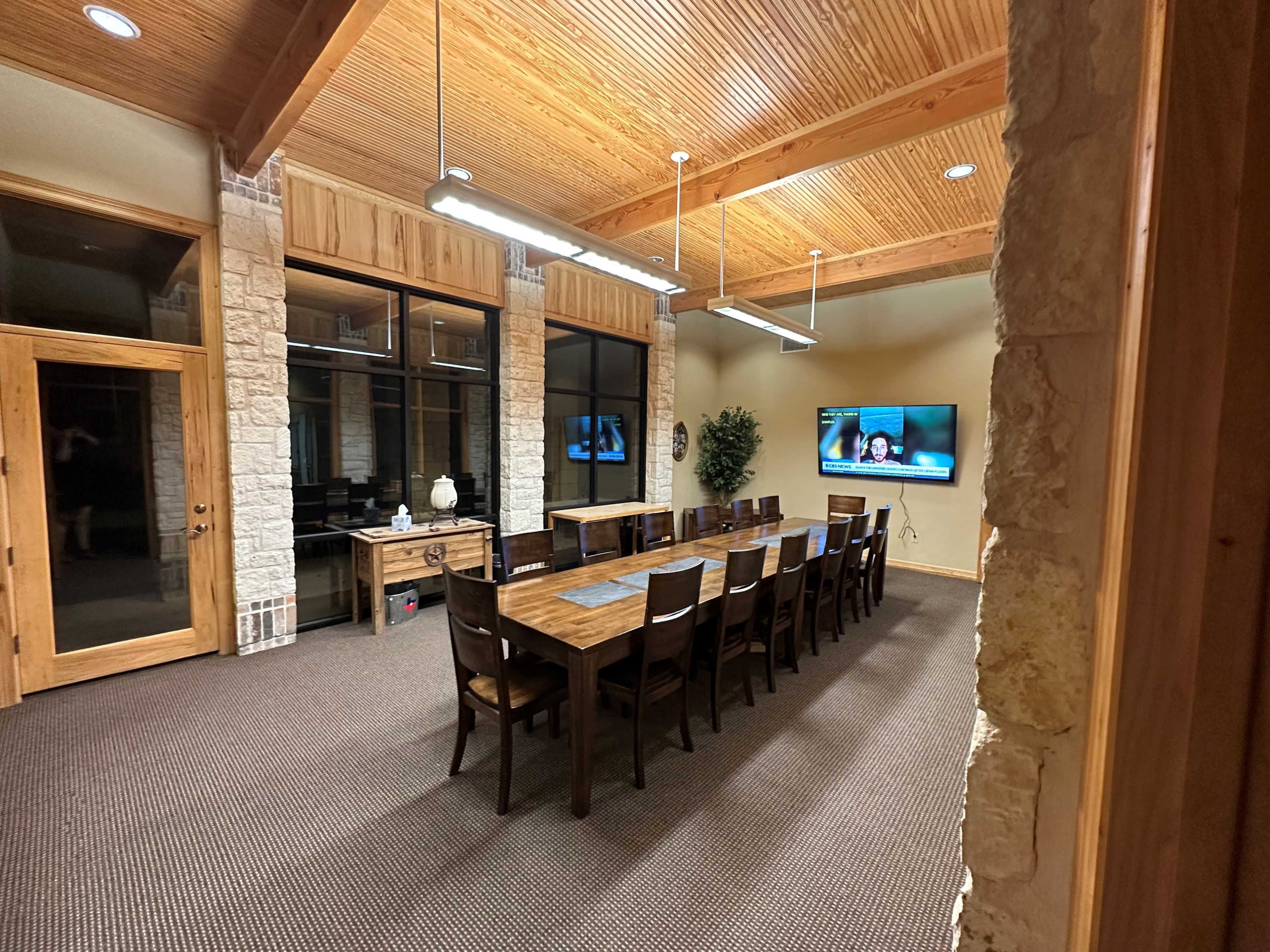 A large conference room with a long wooden table, surrounded by chairs, and a wall-mounted television displaying a video call.