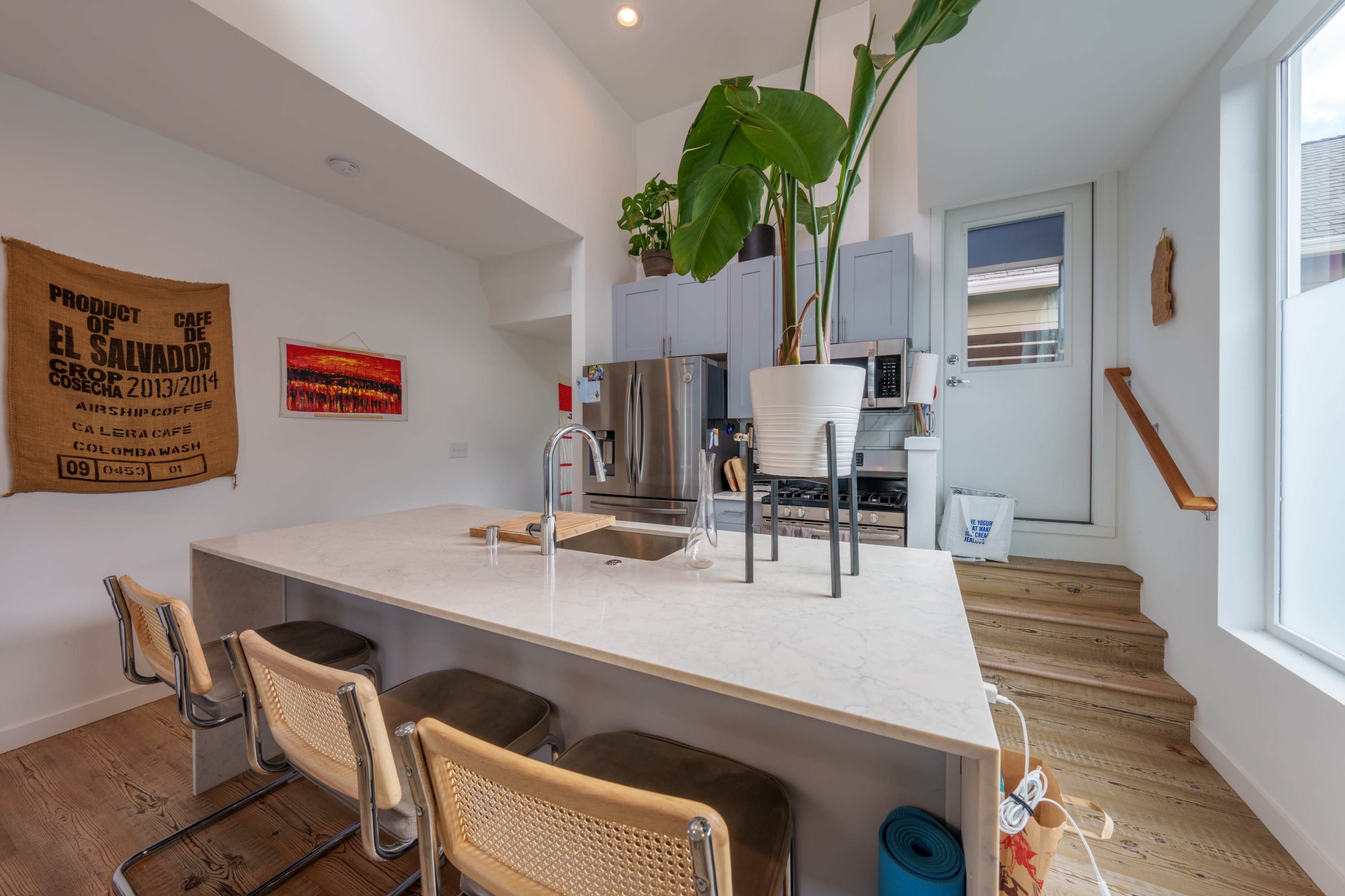 A modern kitchen features a large island with a marble countertop, bar stools, and light cabinetry, along with a potted plant beside a staircase and a window.