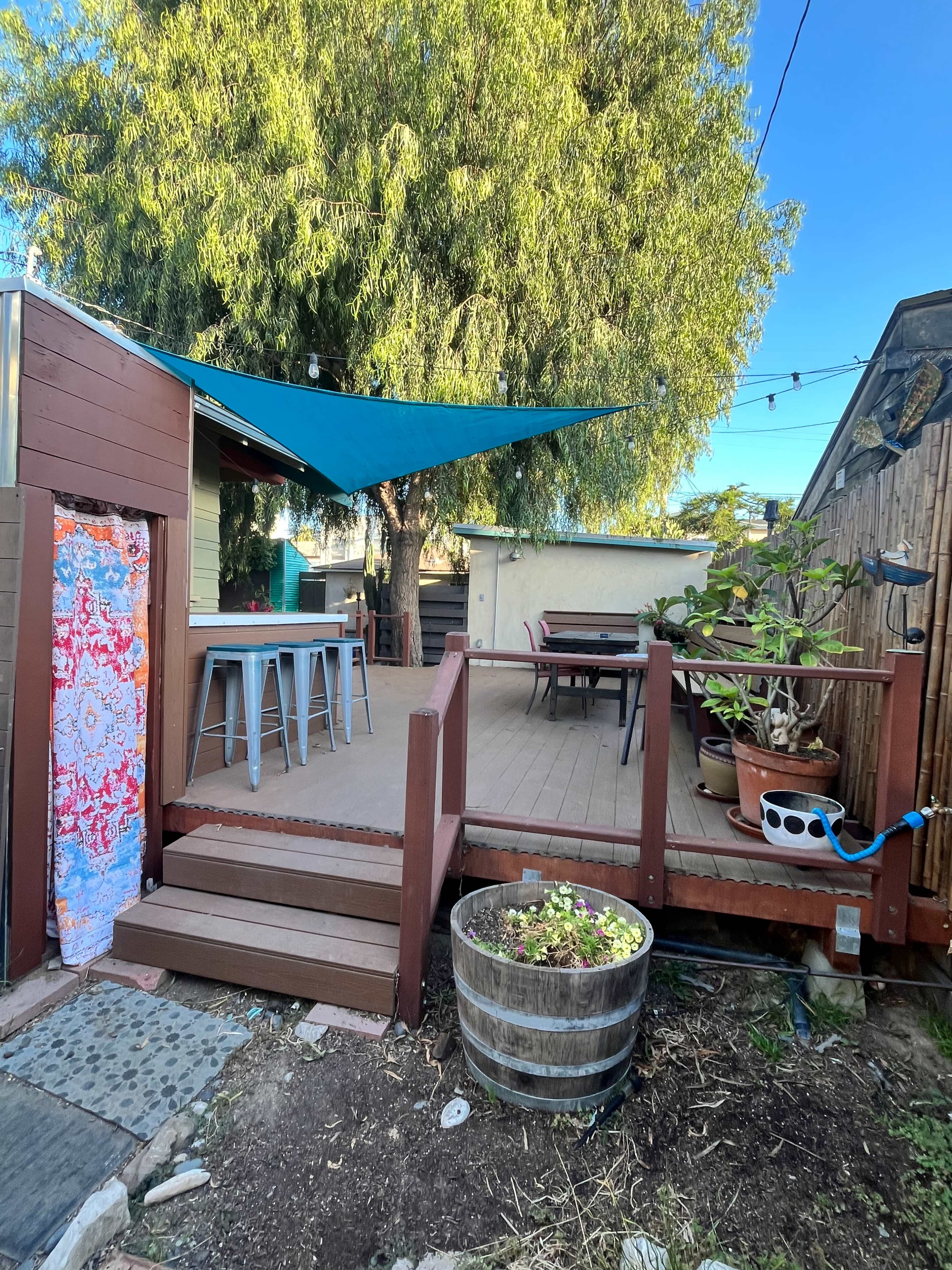 The image shows a wooden deck with a blue shade sail, surrounded by potted plants and steps leading to the garden area.