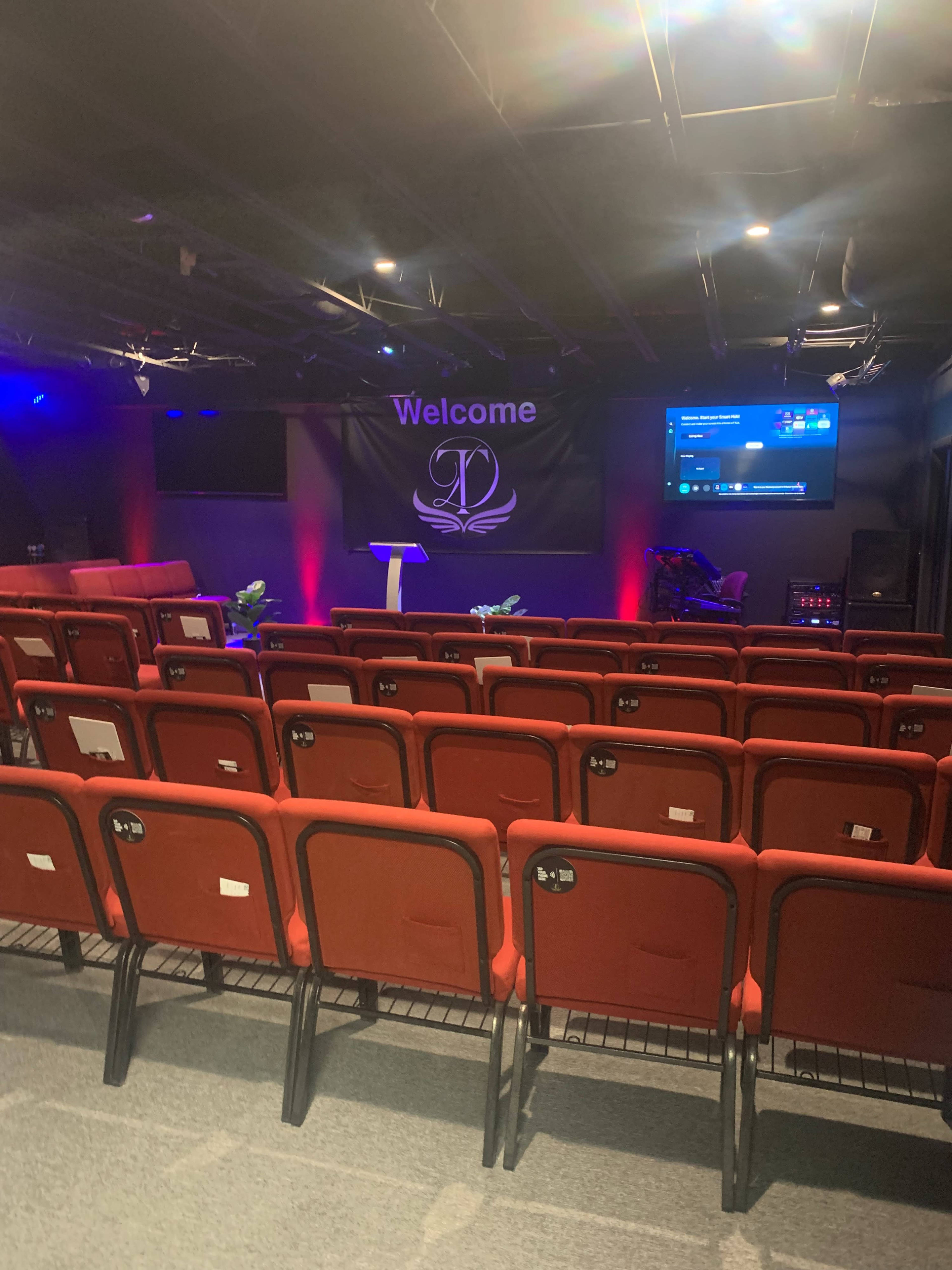 The image shows a small auditorium with red seating arranged in rows, a podium at the front, and a screen displaying a welcome message.