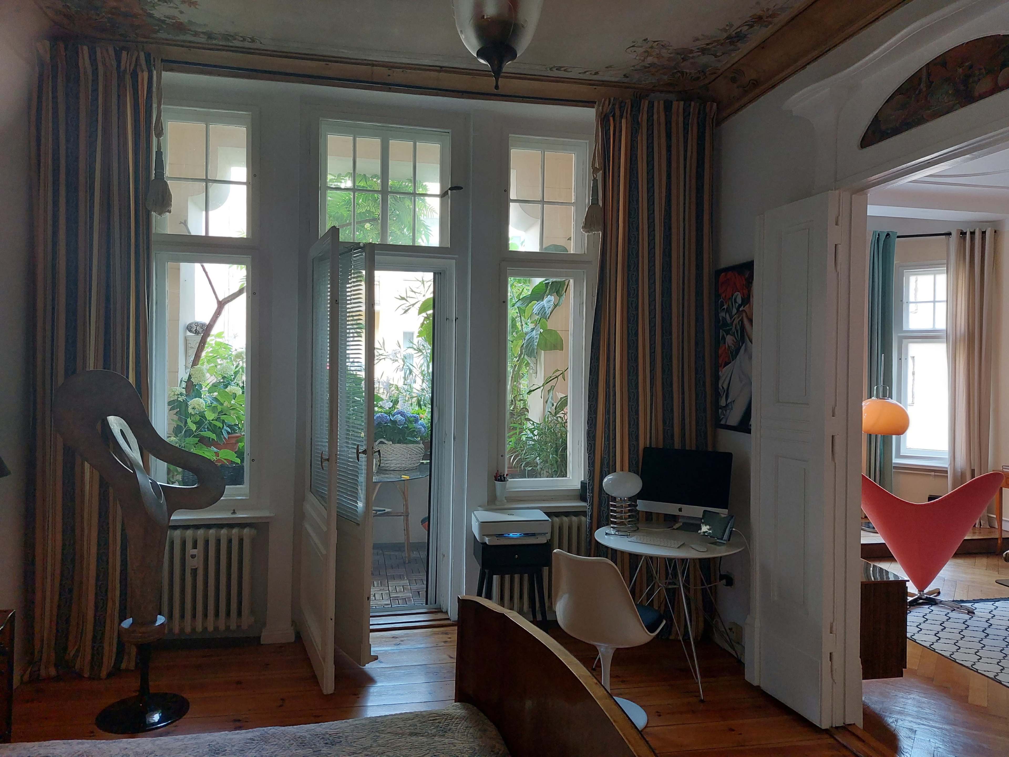 A brightly lit room featuring a mix of modern furniture, large windows with plants outside, and a blend of decorative elements.