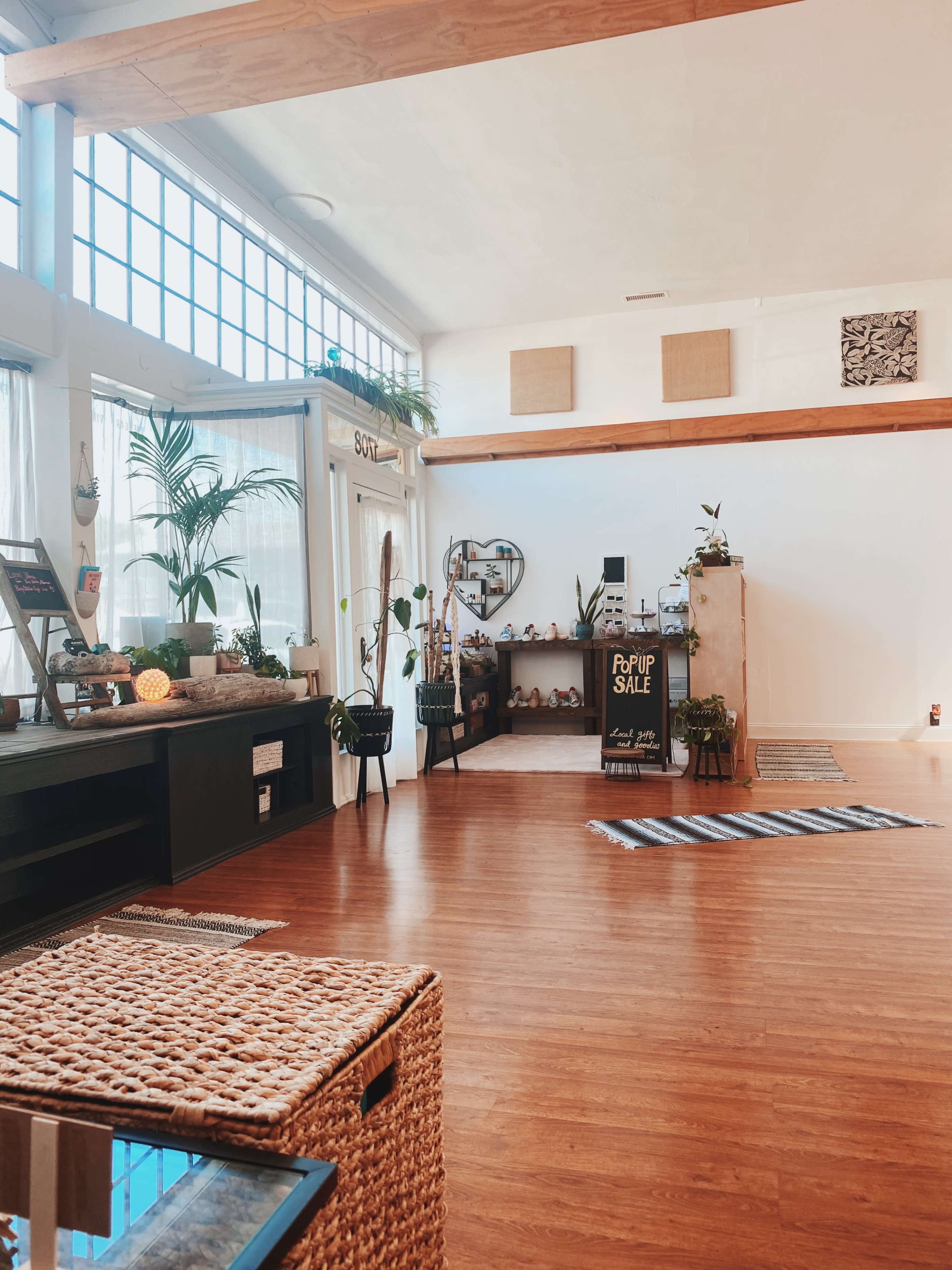 A spacious, well-lit room with wooden floors, large windows, and various plants and decor items arranged around a central display for a pop-up sale.