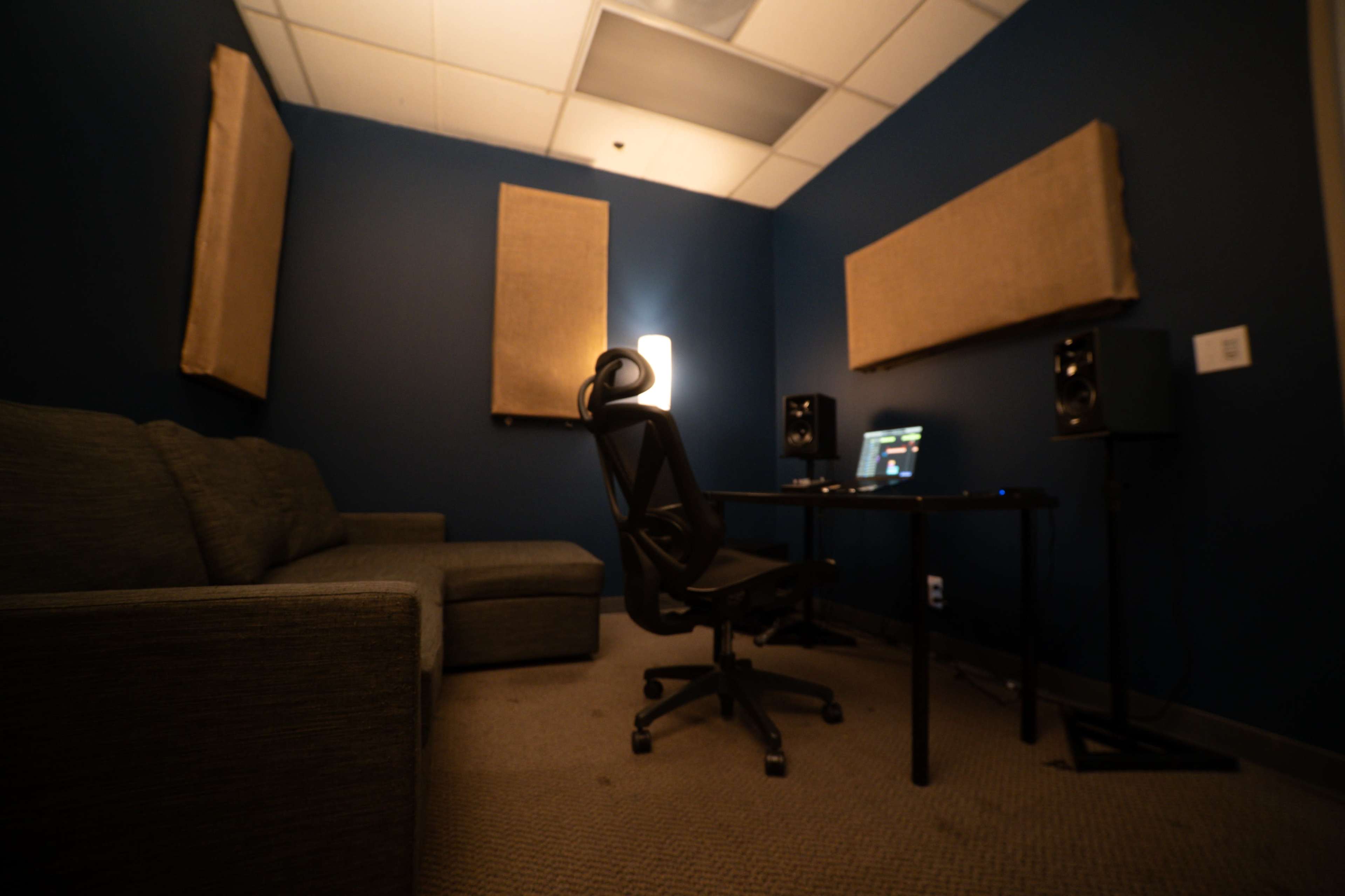 The image shows a small recording studio with soundproofing panels on the walls, a desk with computer equipment, a chair, and a couch in the corner.