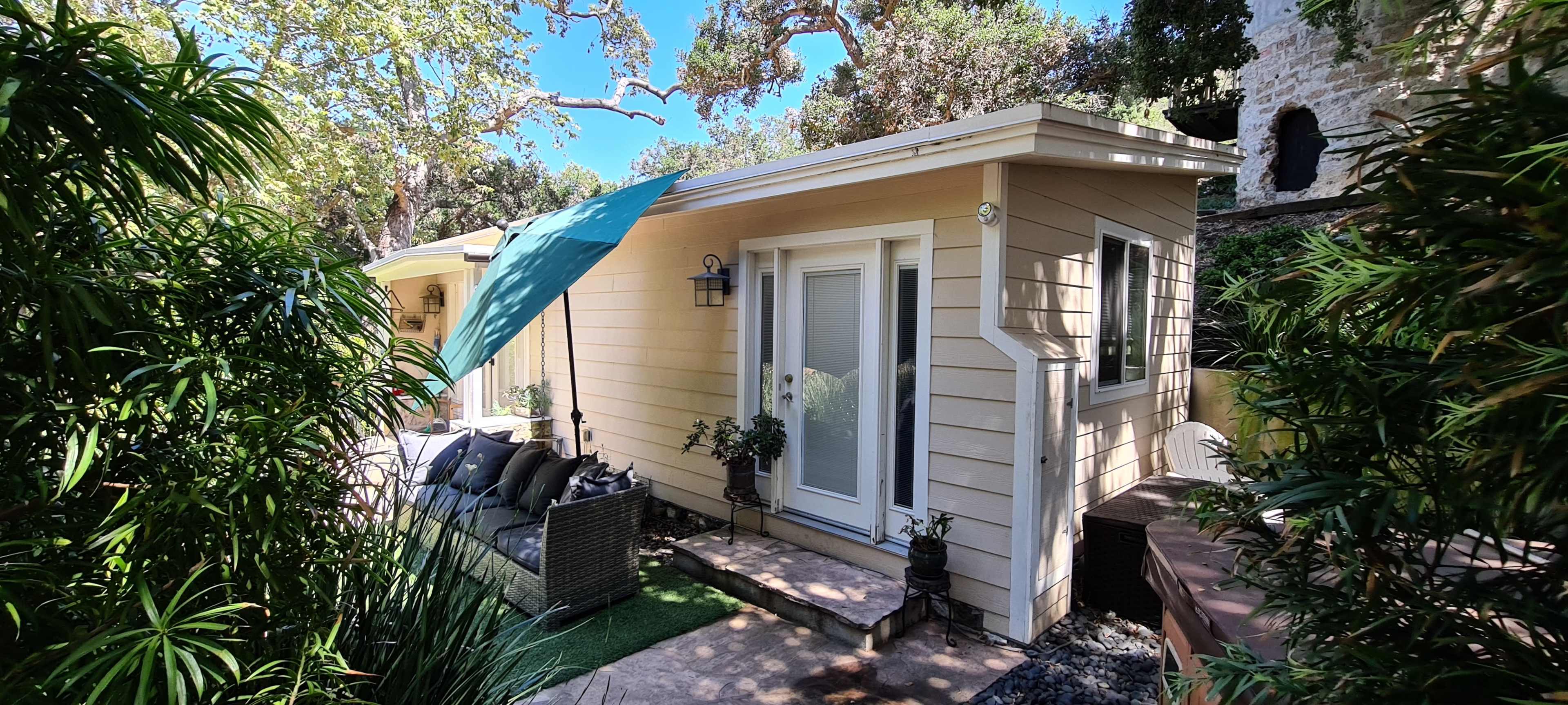 A small yellow cottage with a porch is surrounded by greenery and features an outdoor seating area.