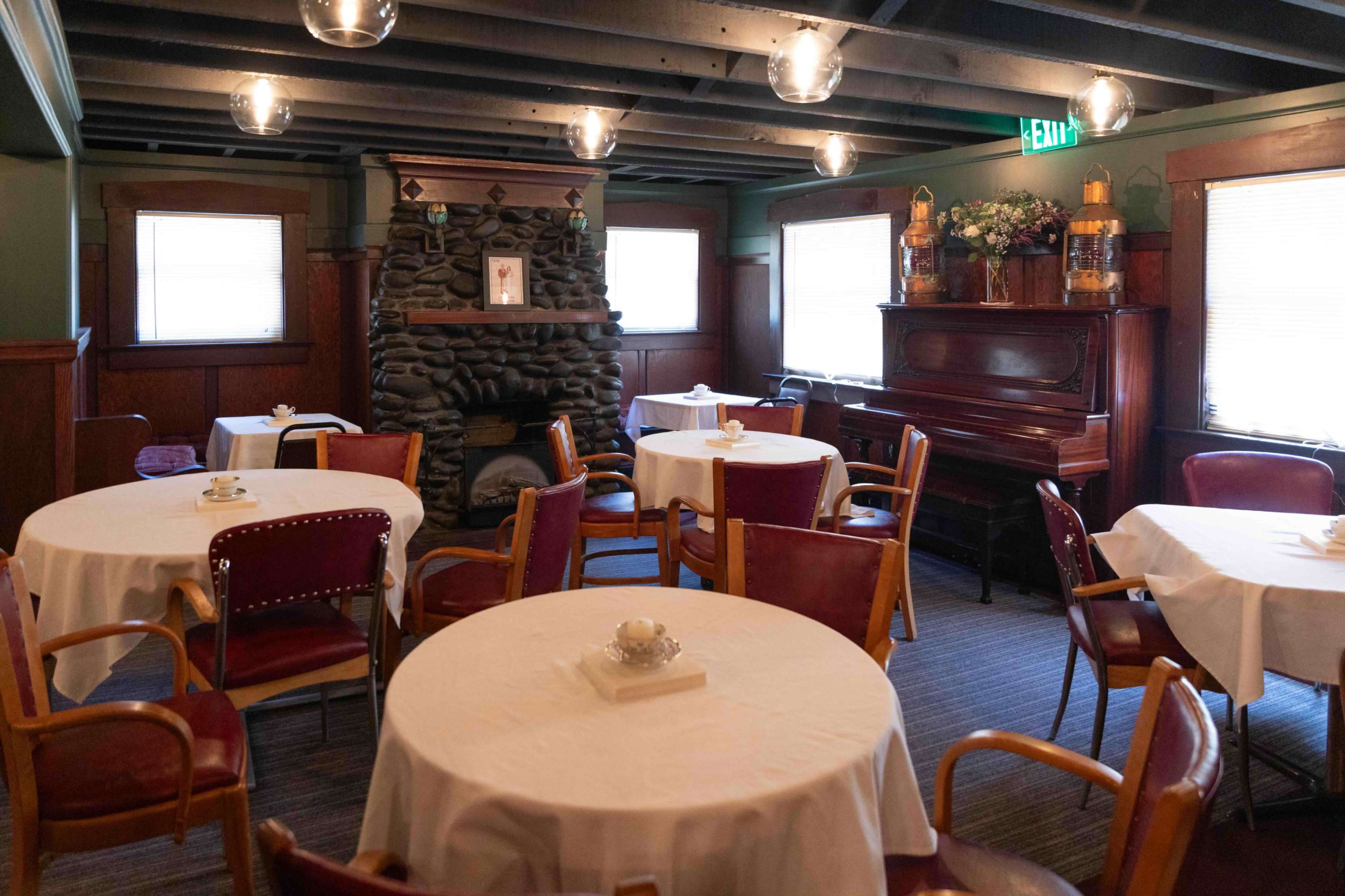 The interior of a cozy restaurant features round tables with white tablecloths, wooden chairs, a stone fireplace, and a piano against a dark-paneled wall.