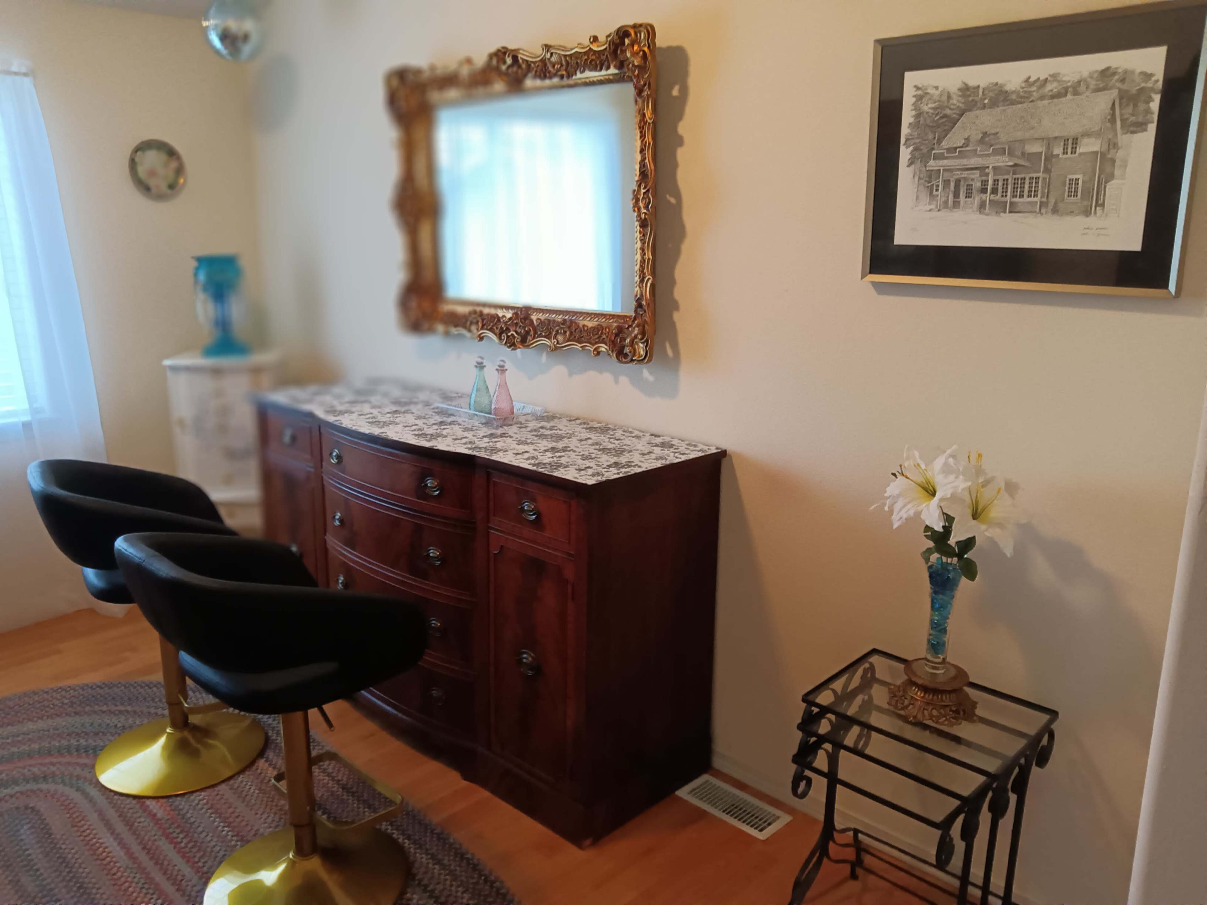 The image shows a cozy room with a wooden dresser topped with a patterned surface, two bar stools, a decorative mirror, a sketch in a frame, and a vase with flowers on a side table.