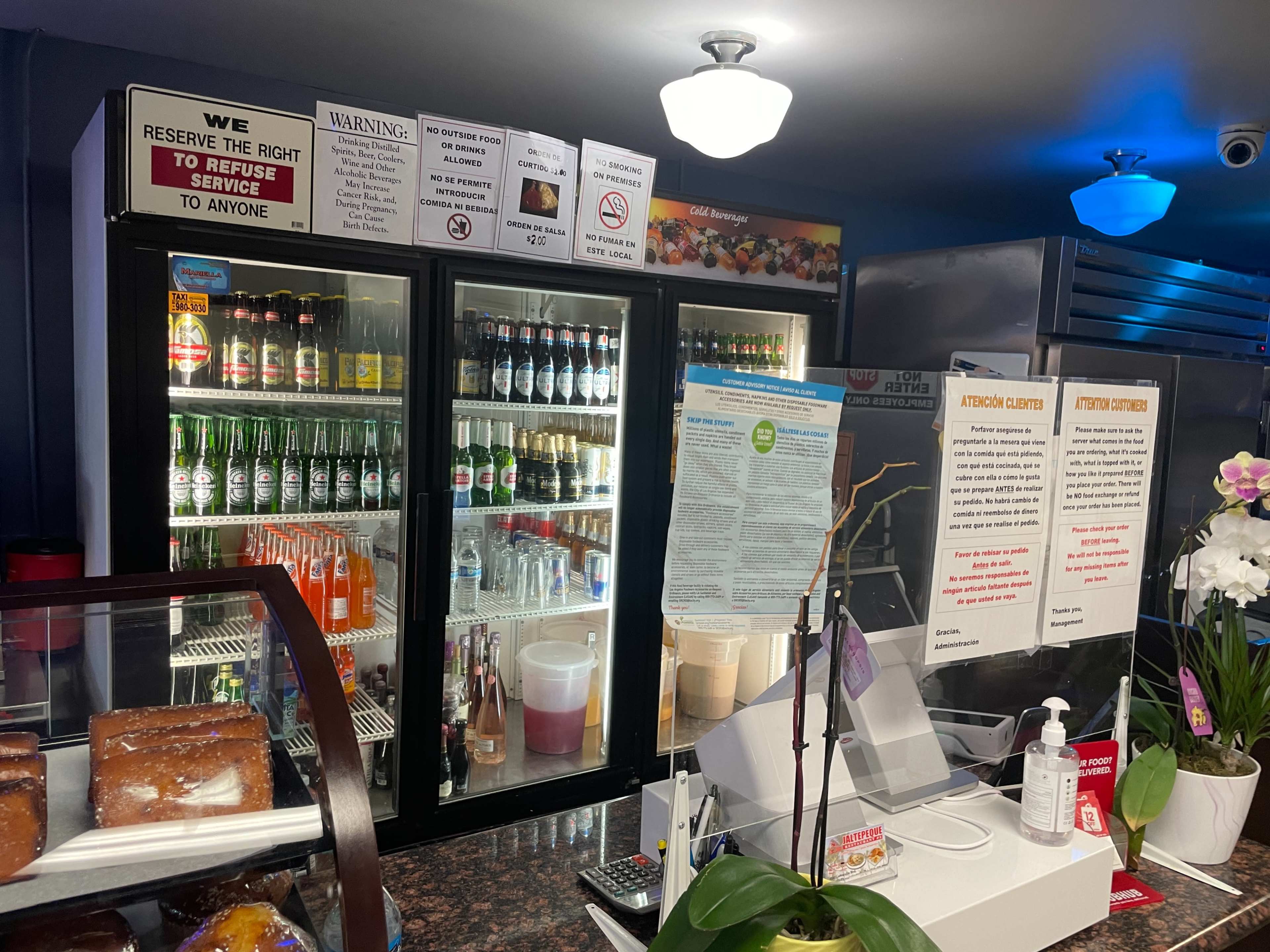 A service counter in a restaurant featuring a display of beverages in glass-front refrigerators, snacks under a glass case, and various signs about service policies.