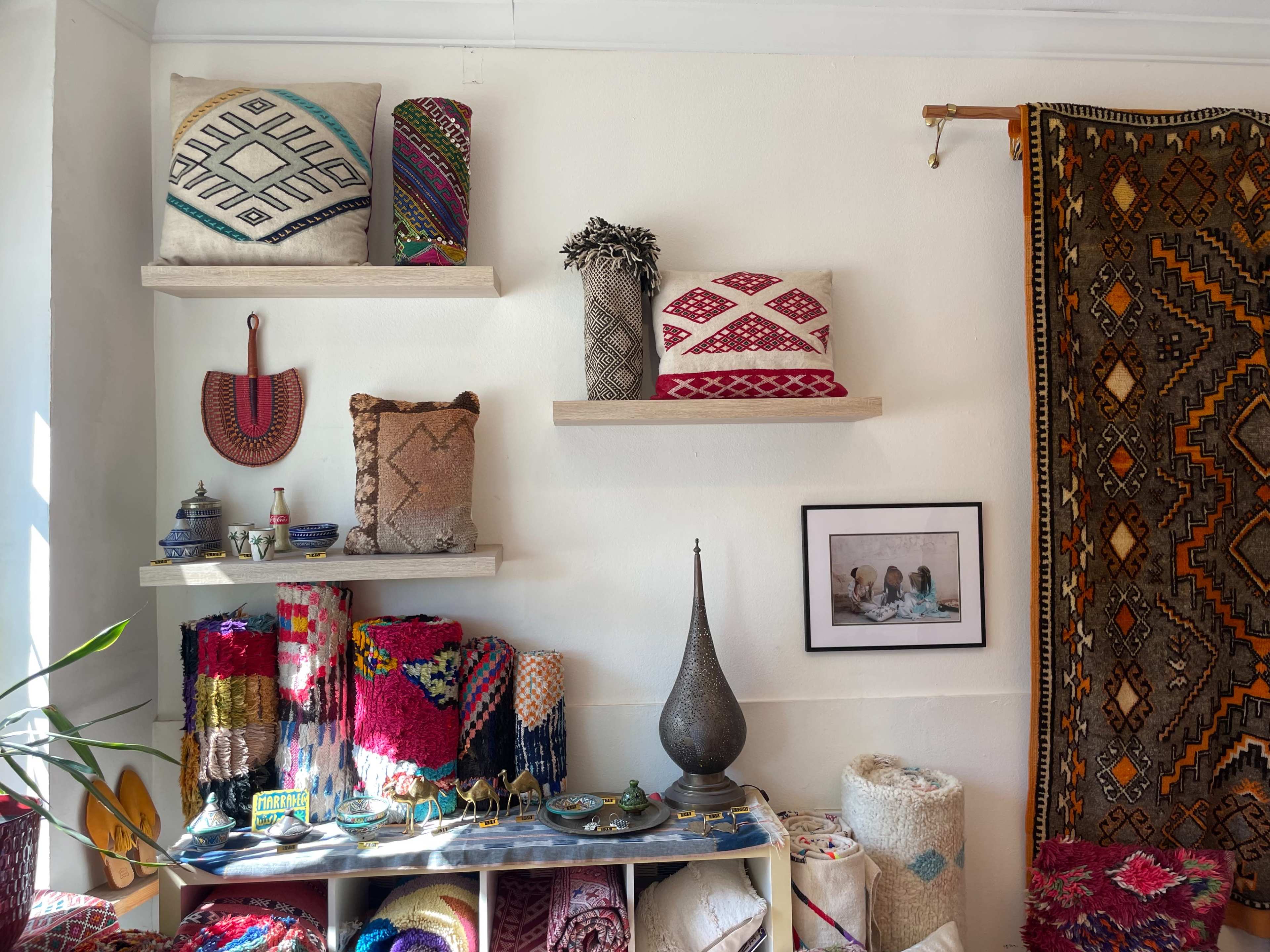 The image shows a cozy interior filled with colorful textiles, decorative pillows on shelves, and various handcrafted items arranged on a table.