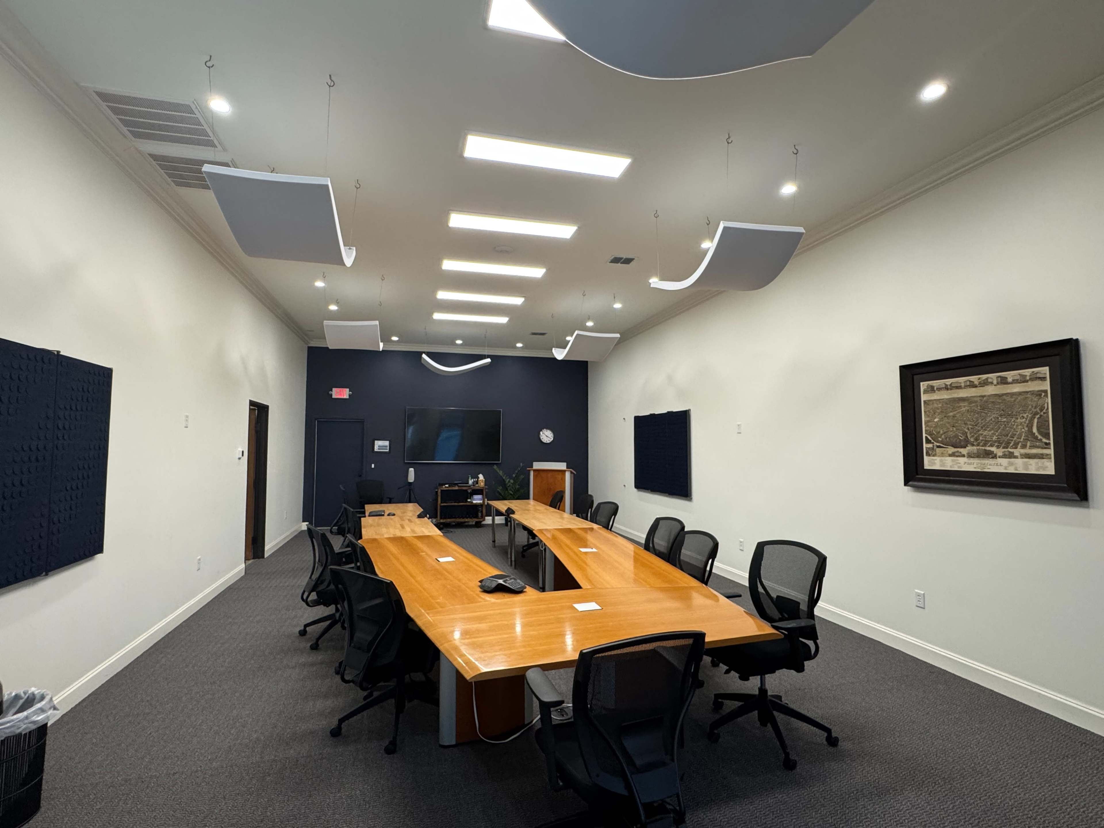 The image depicts a modern conference room featuring a long wooden table surrounded by black office chairs, with acoustic panels on the walls and a large screen at one end.