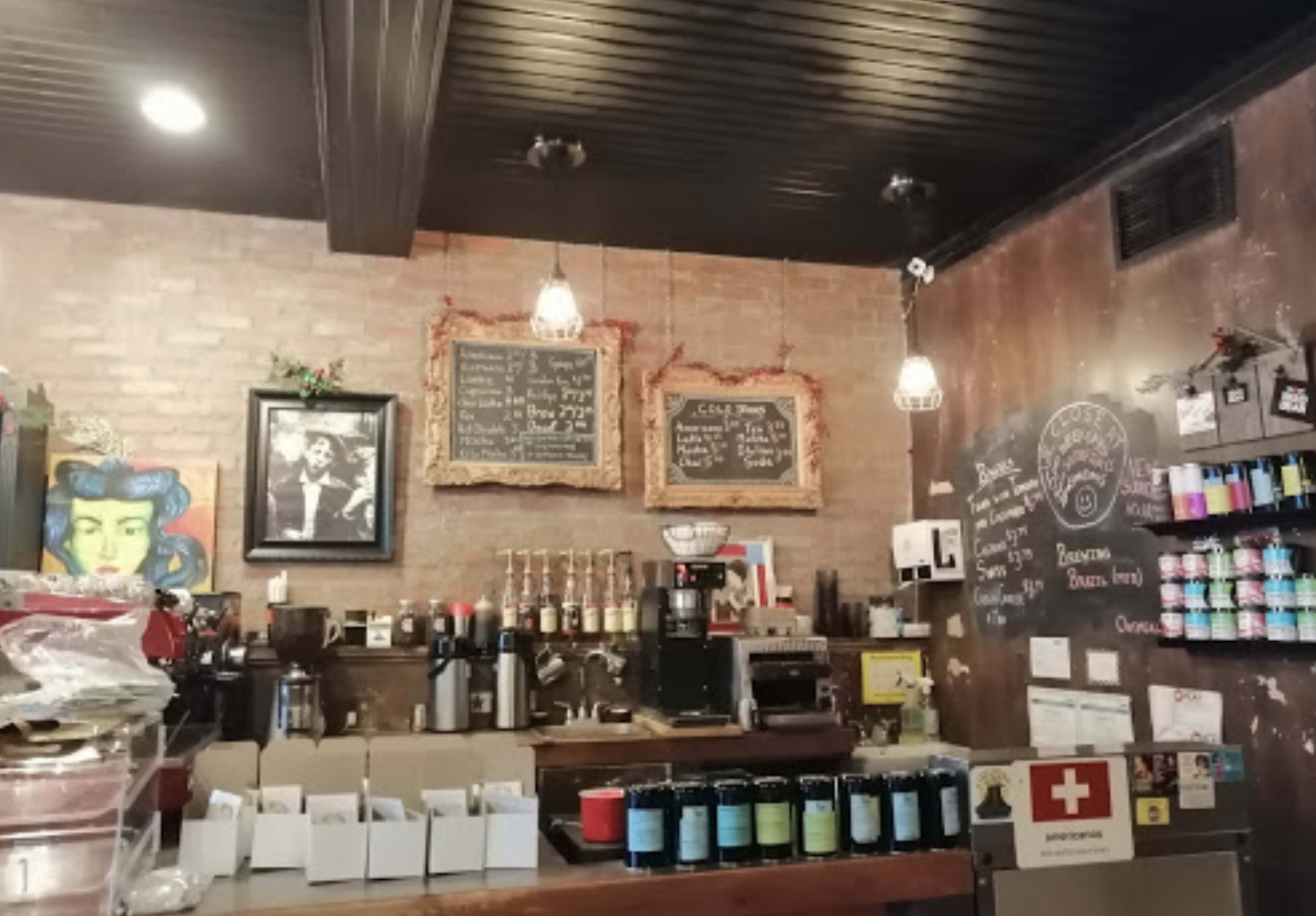 The interior of a coffee shop features a wooden counter with various coffee equipment, a chalkboard menu on the wall, and framed art pieces.