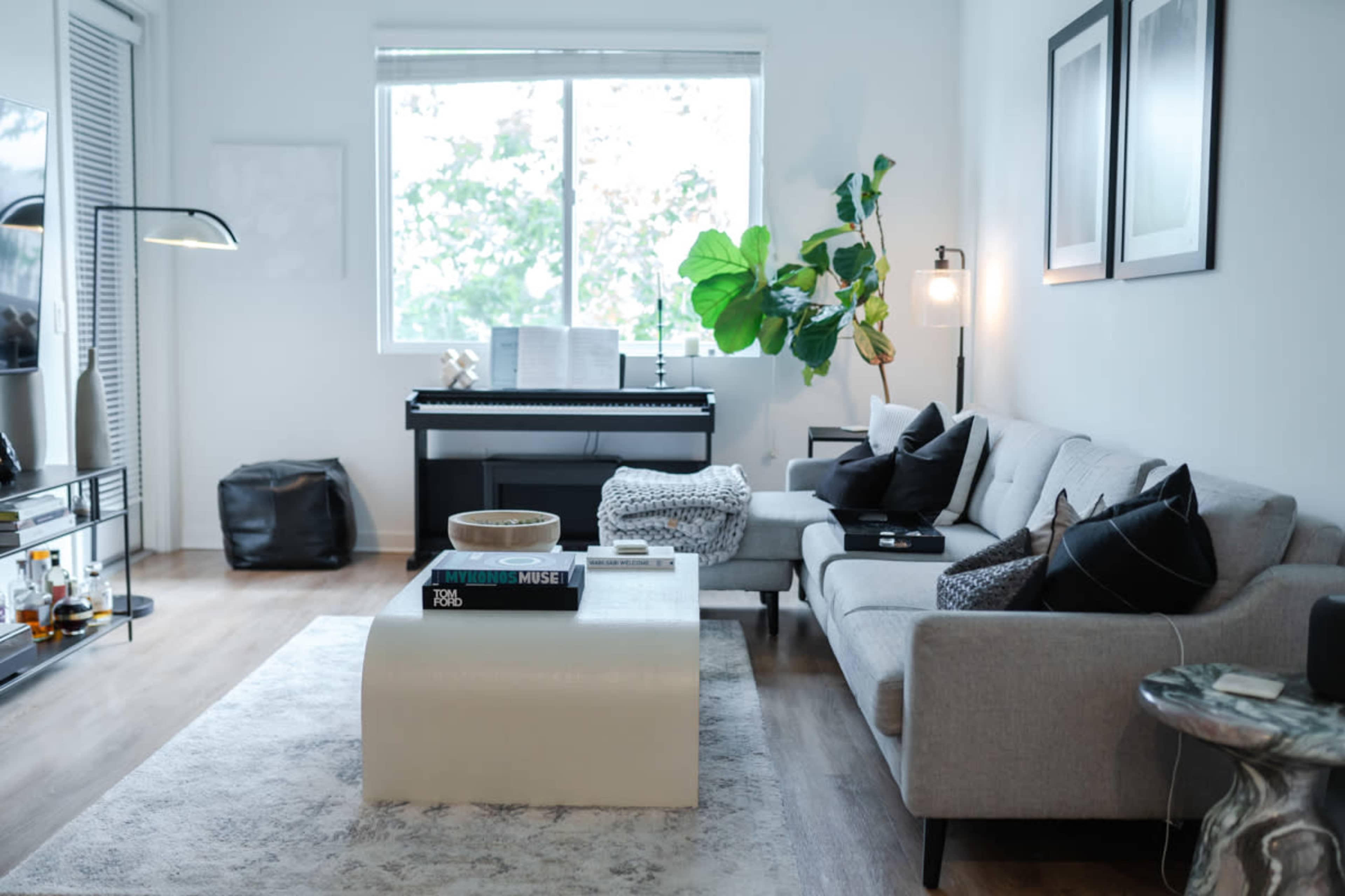 A modern living room features a light gray sectional sofa, a coffee table, and a digital piano near a large window, illuminated by natural light.