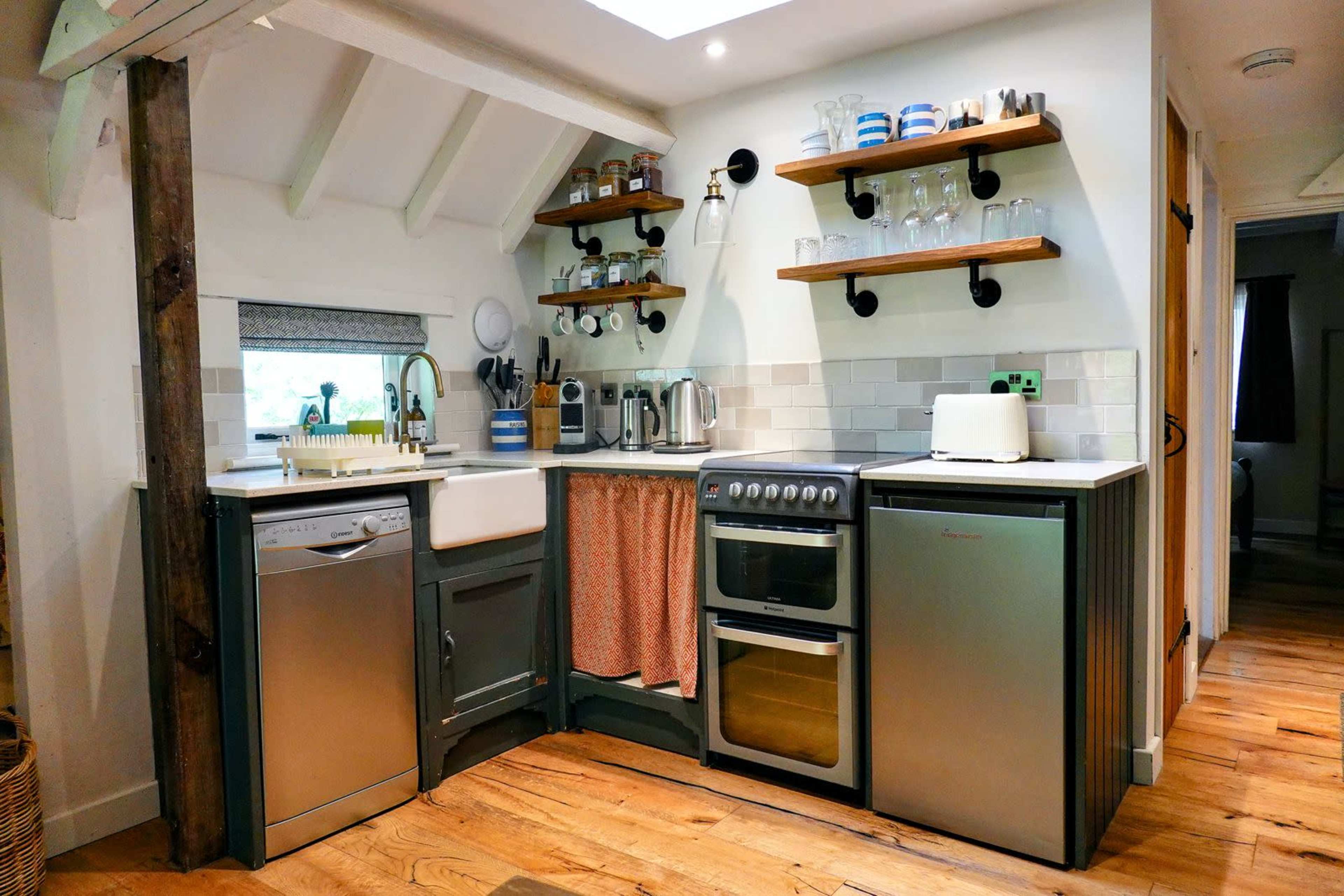 A compact kitchen features a sink, stove, dishwasher, and shelves with glassware and dishes, all arranged against a wooden floor and light-colored walls.