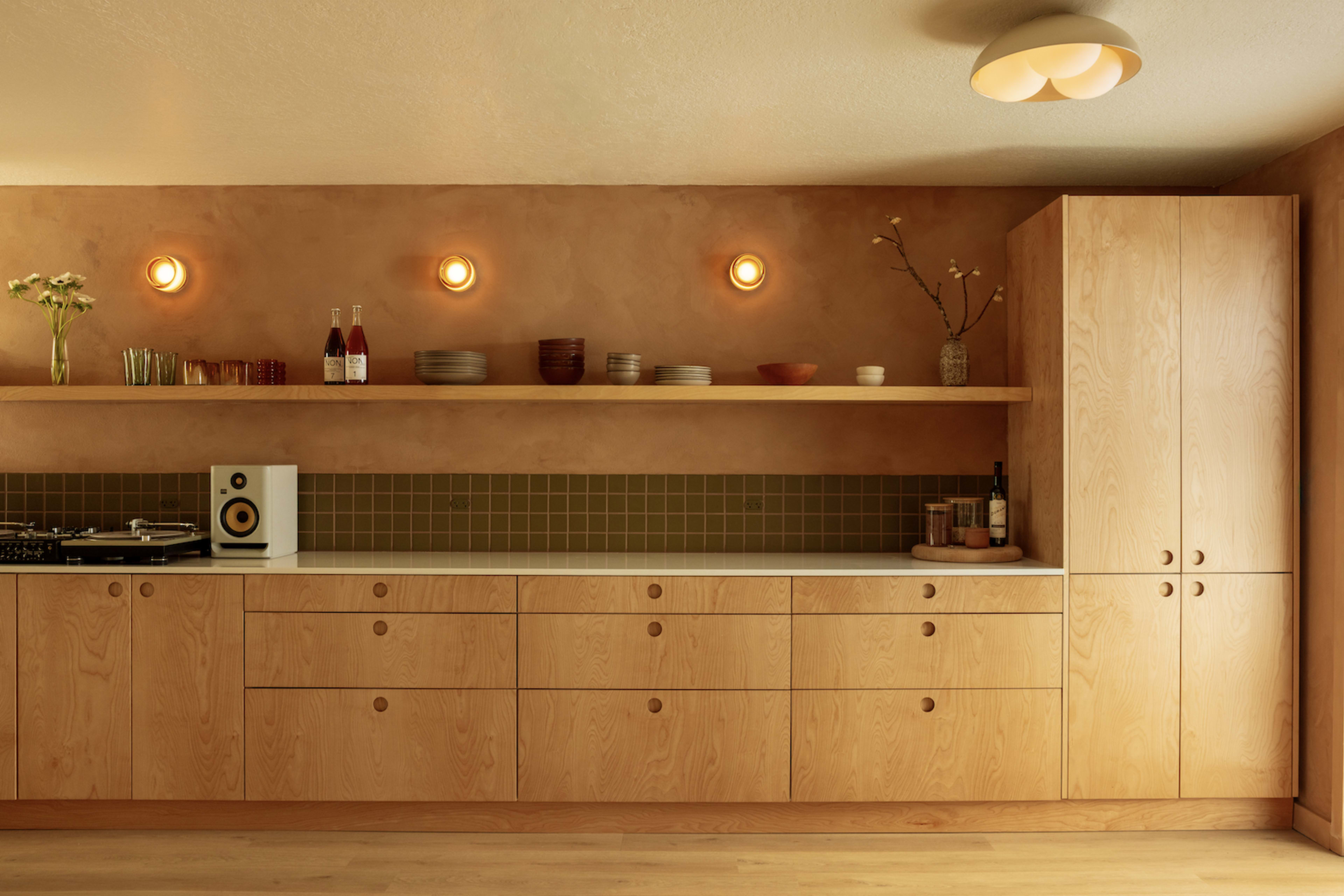 The image shows a modern kitchen with light wooden cabinetry, a white countertop, and a green backsplash, featuring shelves with decorative items and two bottles.