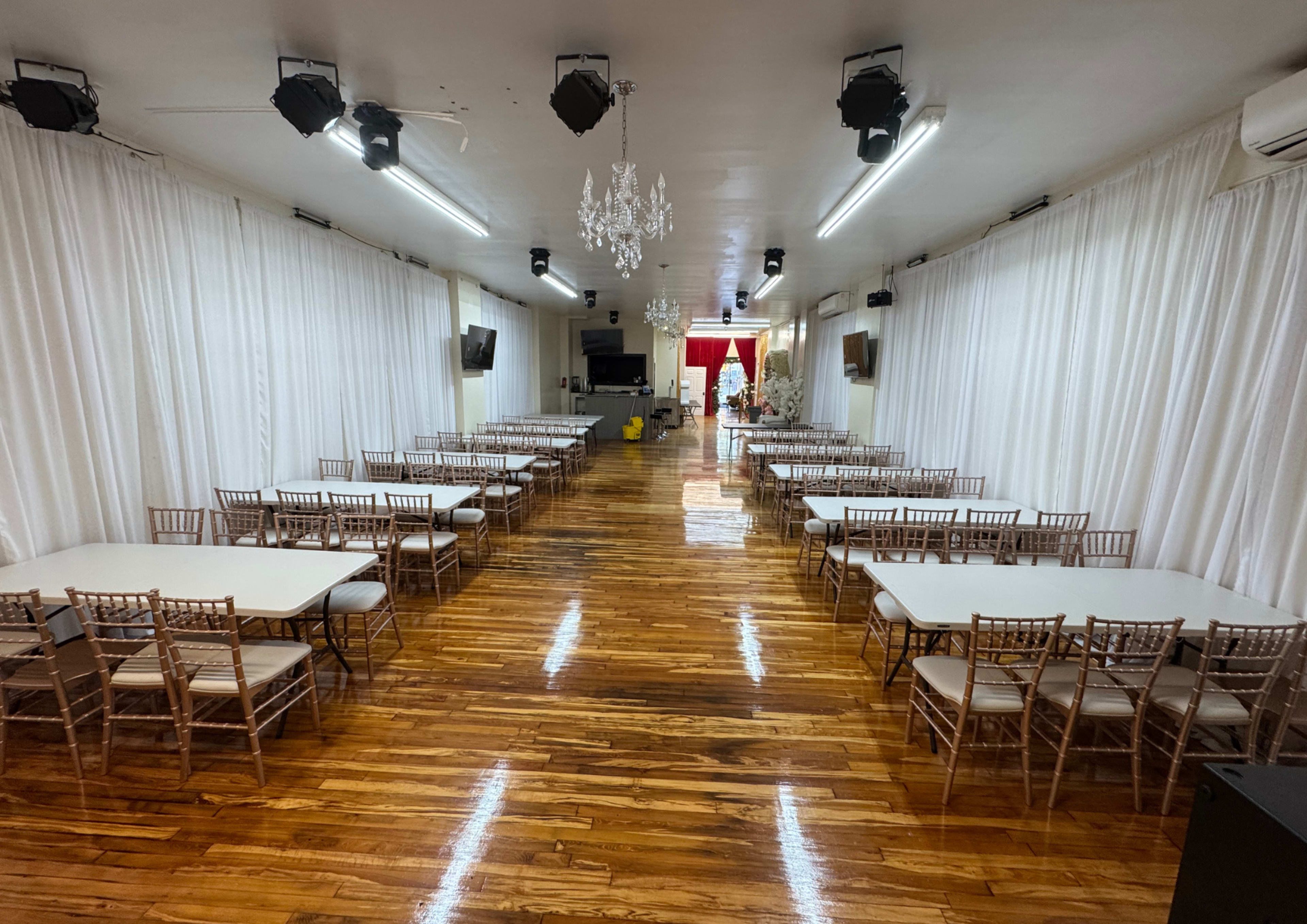 The image shows a large event space with polished wooden floors, white draped walls, rows of tables surrounded by chairs, and a decorative chandelier hanging from the ceiling.