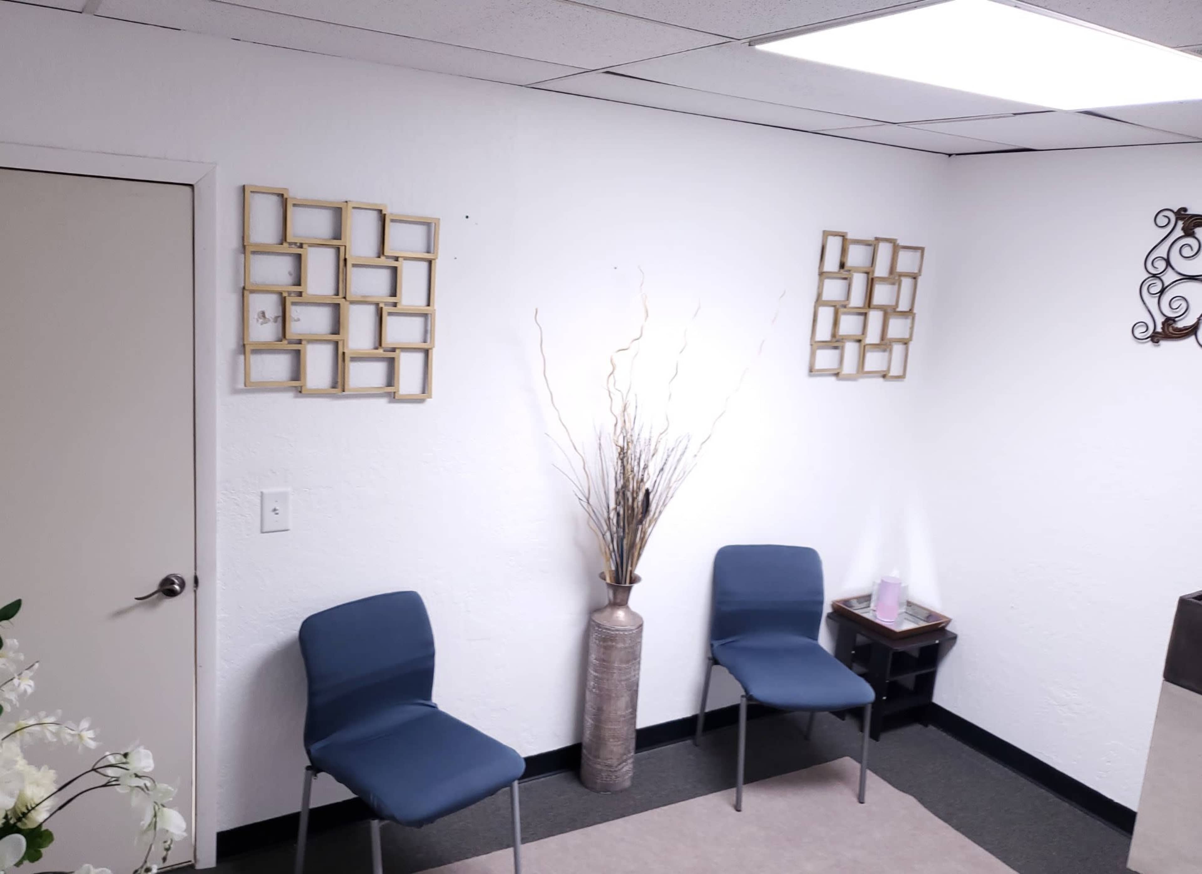 A waiting area with two blue chairs, a vase with branches, and decorative frames on the white walls.